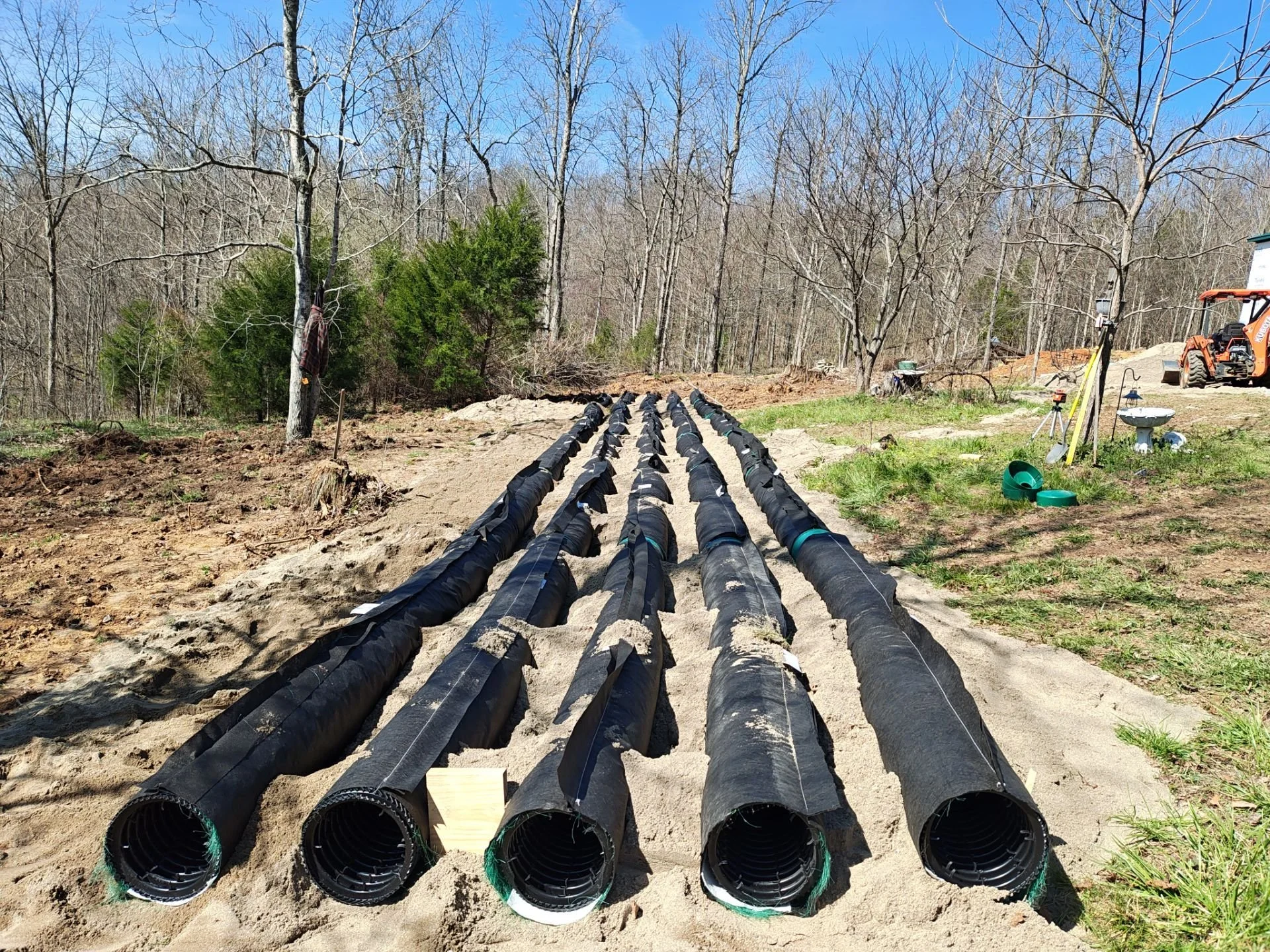 Multiple black pipes laid in a row on a dirt surface in a wooded area, likely for a plumbing or irrigation project, with construction equipment and tools visible in the background.