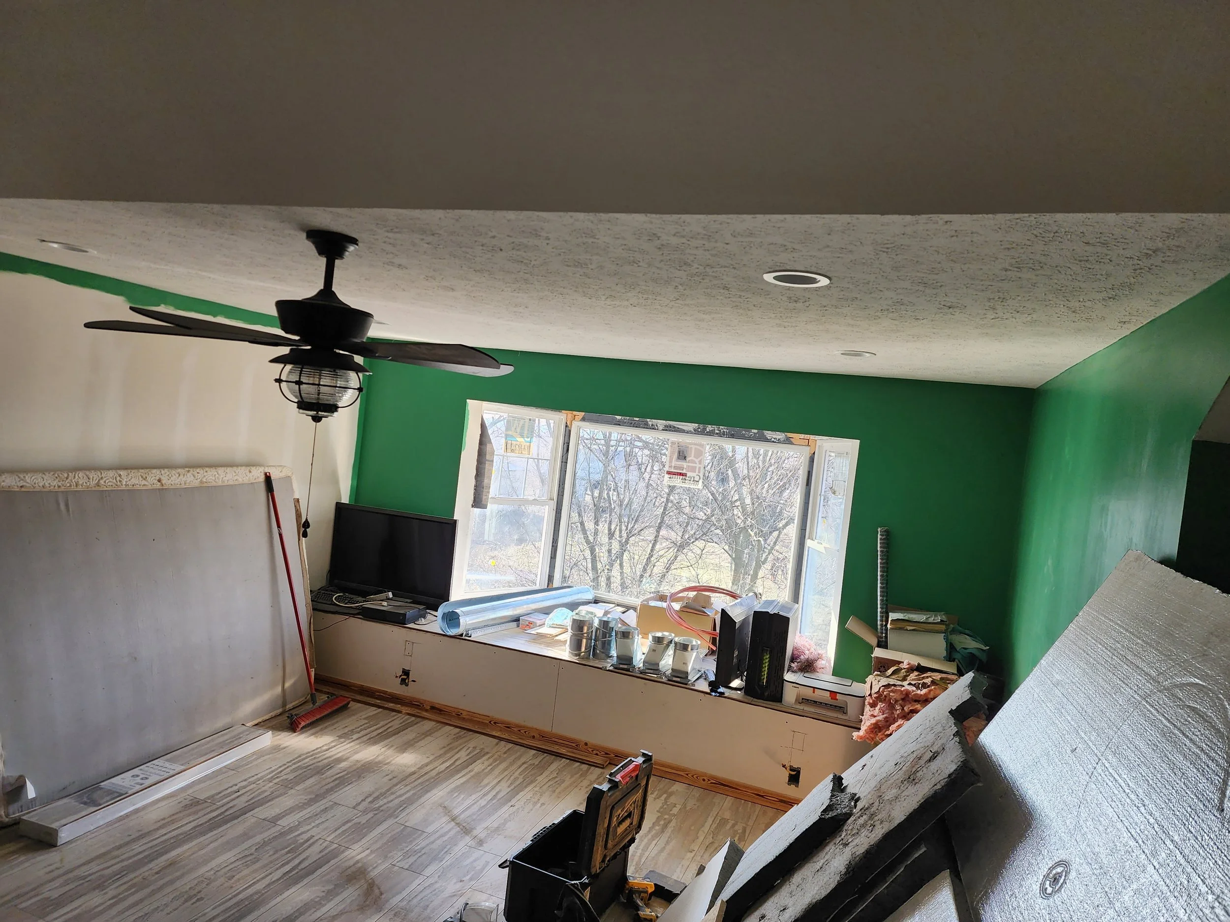 Interior of a room under renovation with green walls, a new ceiling fan, and window providing natural light. Construction tools and materials are scattered around, with a partially installed baseboard and exposed insulation and framing.