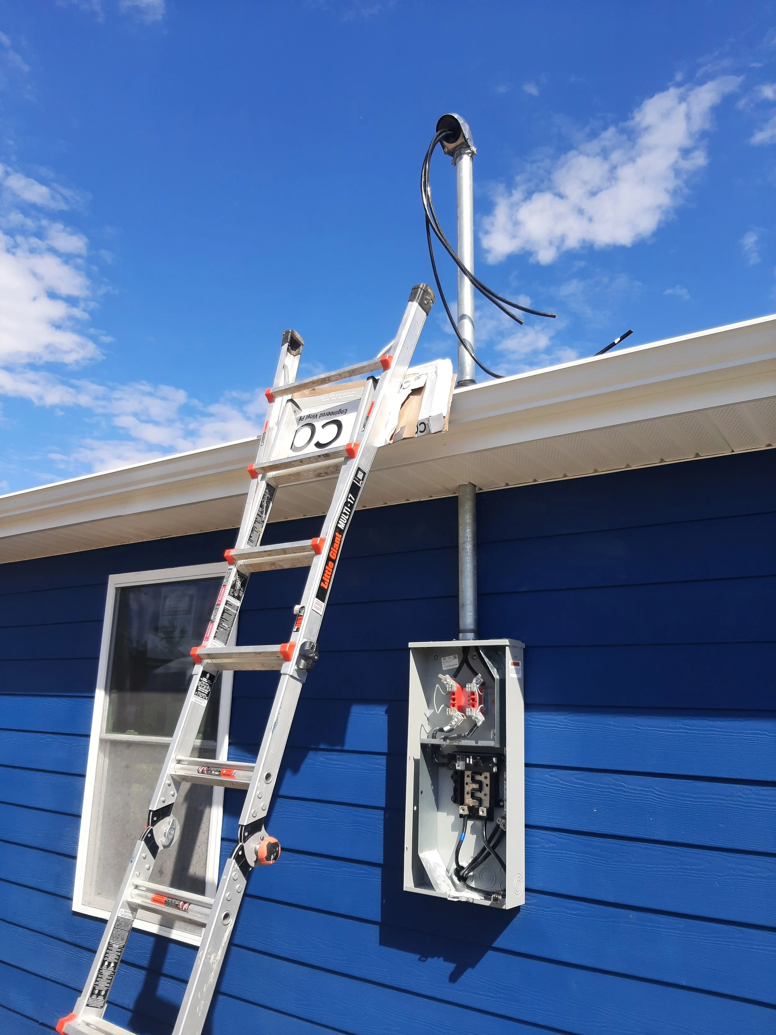 Ladder set up against the side of a blue house under a clear sky, with an electrical box and a conduit pipe on the wall, and a weatherproof outlet on the ladder.
