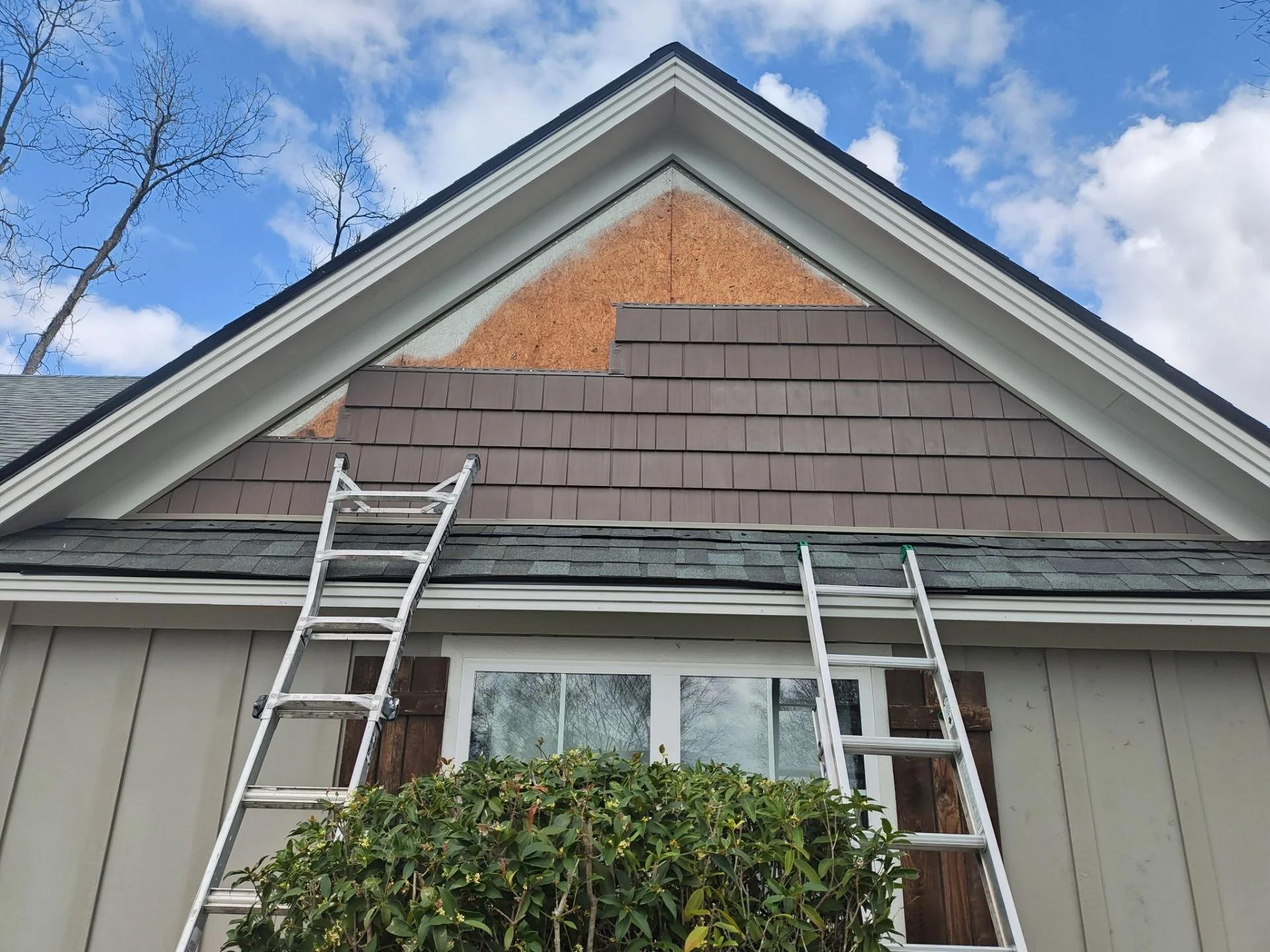 Exterior of a house with a gable roof, showing the soffit and fascia. Two ladders are leaning against the house, with a bush in front and a window below. The upper part of the house is under renovation, exposing the sheathing and insulation underneath the siding.