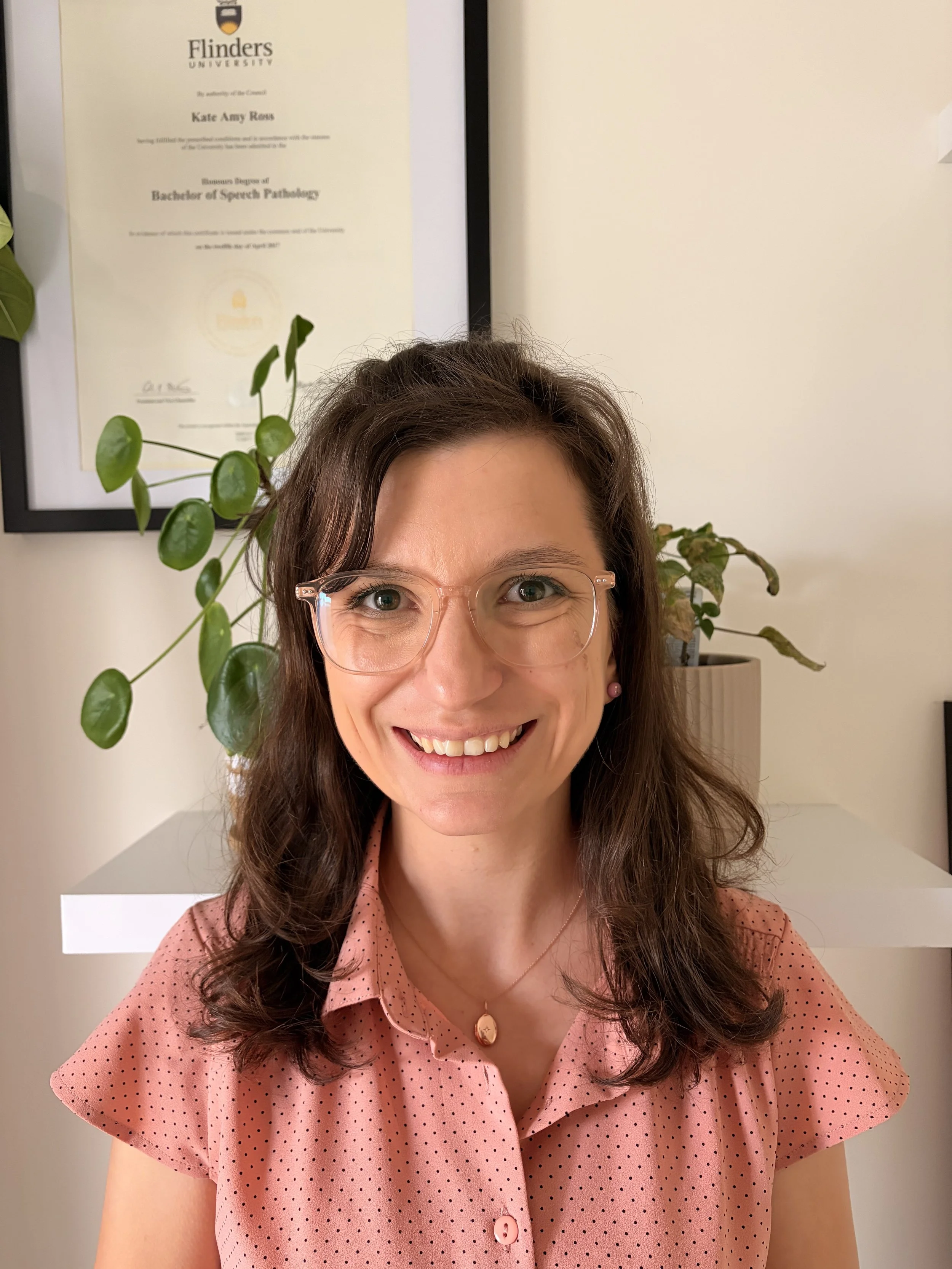 Kate Ross Speech Pathologist. Portrait of a woman with glasses smiling, wearing a pink polka dot blouse, with a framed certificate on the wall and a potted plant behind her.