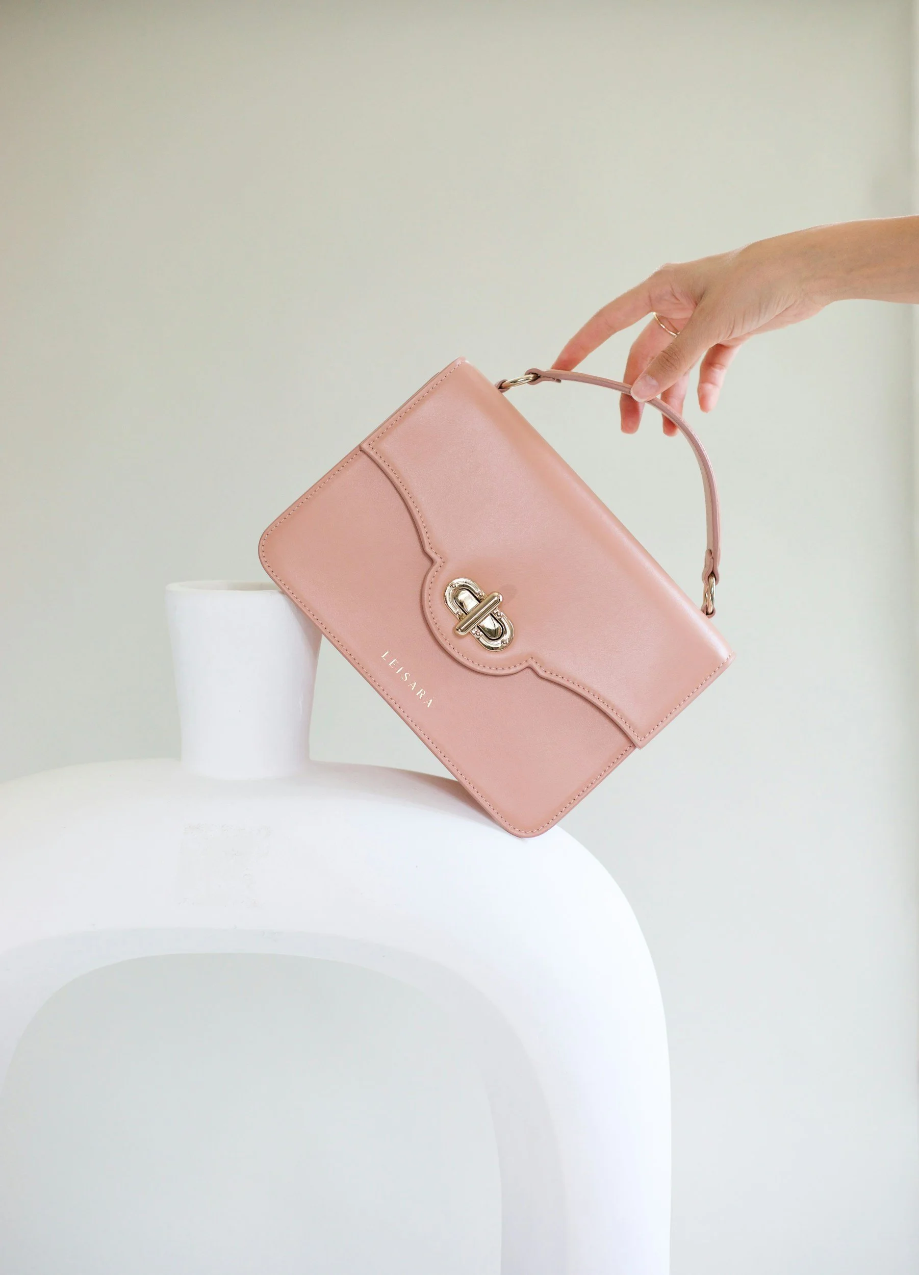A pink handbag with a gold clasp is being held above a white ceramic vase.