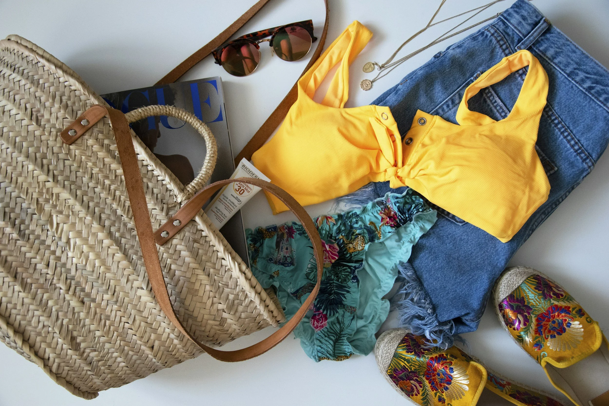 Beach bag with a magazine and sunscreen, with a yellow tank top, turquoise patterned swim shorts, blue denim jeans, floral espadrille shoes, sunglasses, and jewelry laid out nearby.