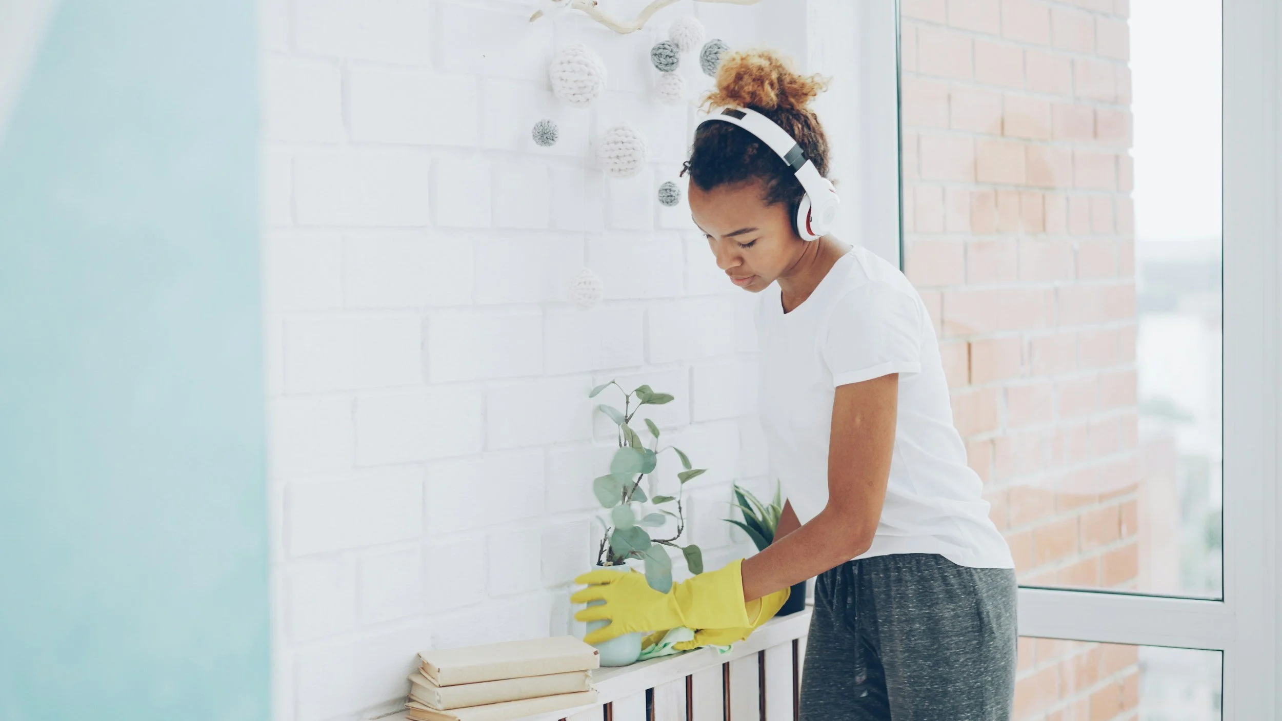 Young woman wearing yellow rubber gloves, white t-shirt, and headphones, cleaning a windowsill with a potted plant and books.