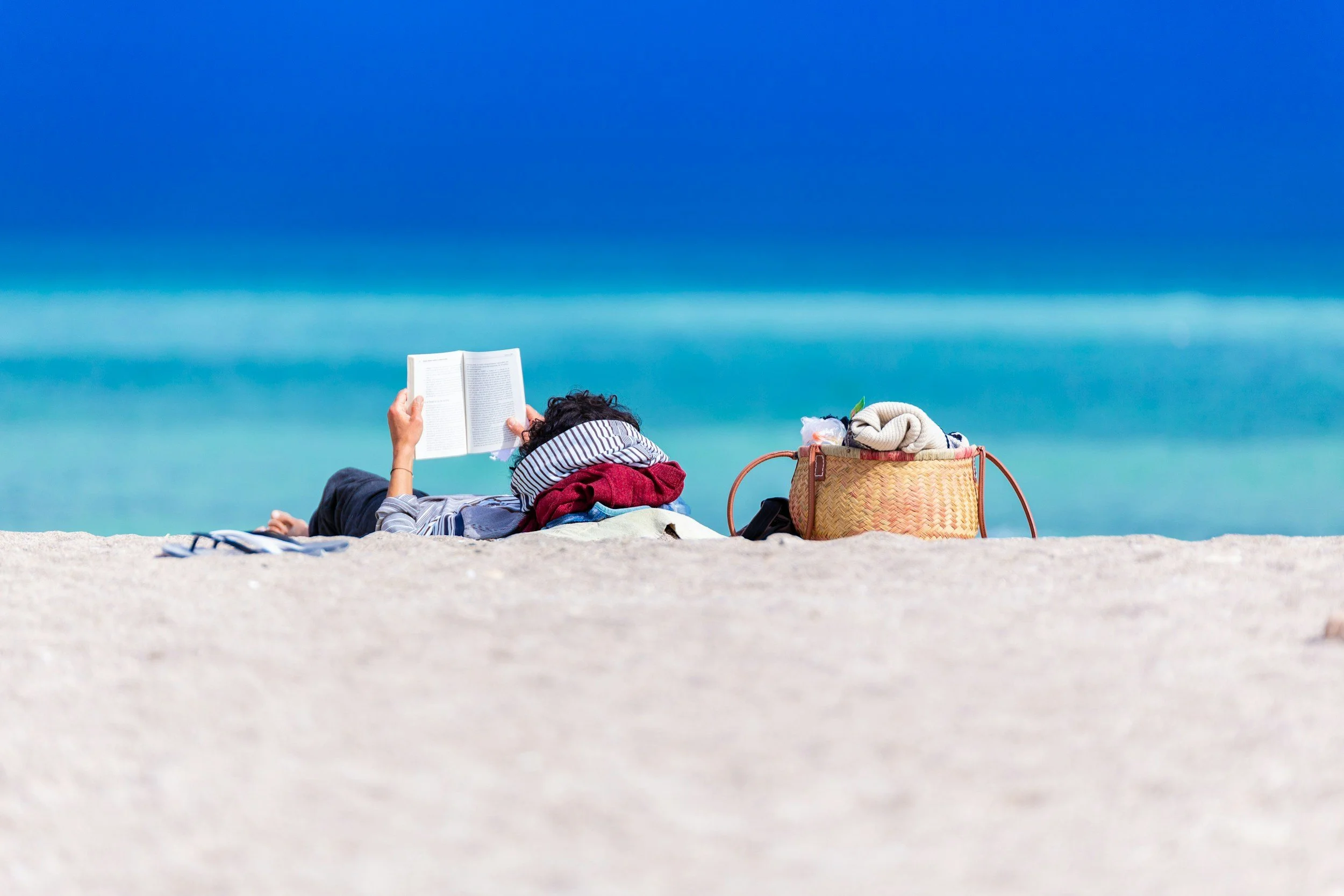 Person lying on the beach reading a book, with a beach bag and clothes nearby, against a clear blue sky and ocean background.