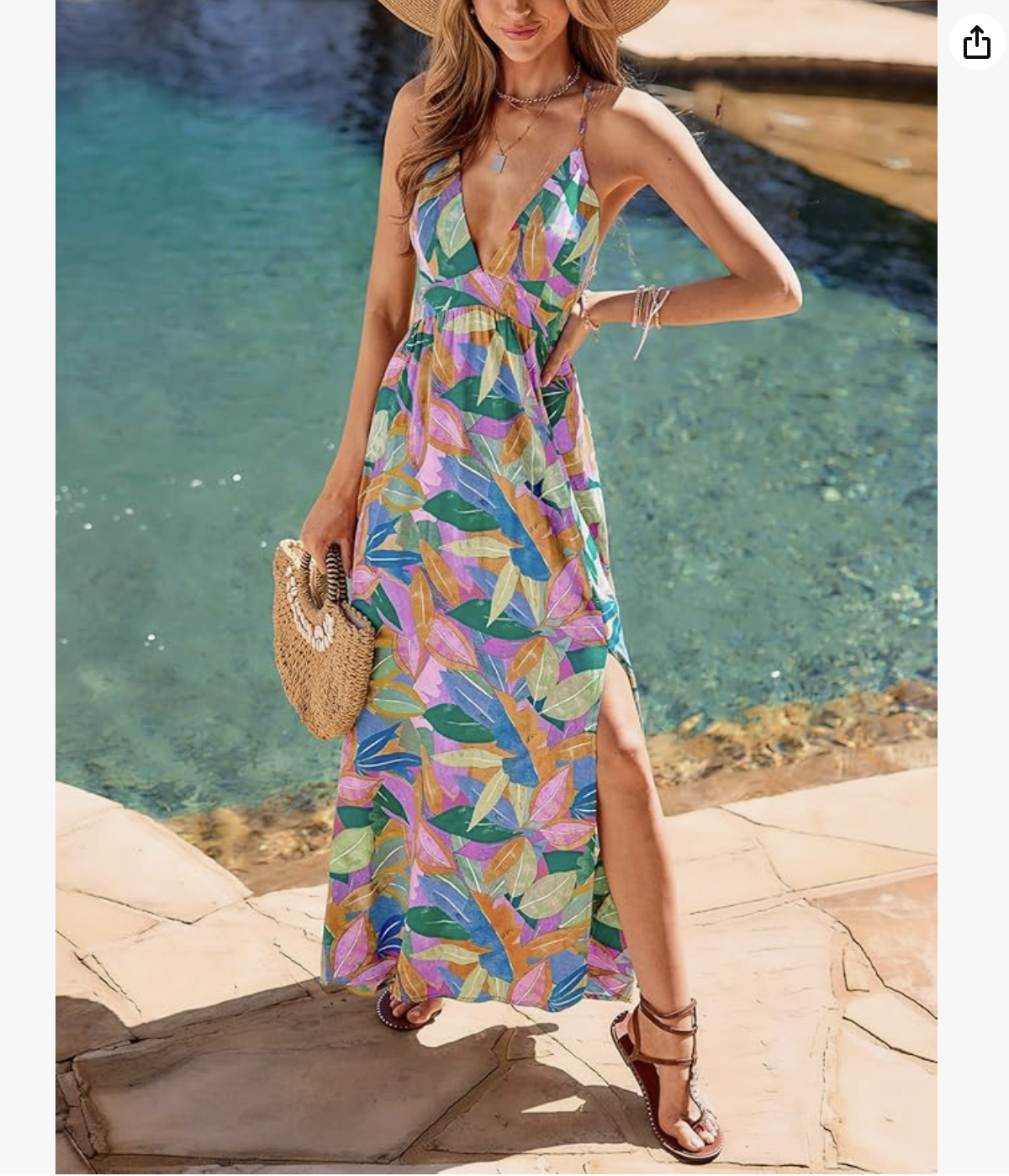 A woman standing by a pool, wearing a colorful, tropical-patterned maxi dress with a slit, accessorized with a straw hat, jewelry, and sandals, holding a woven purse.