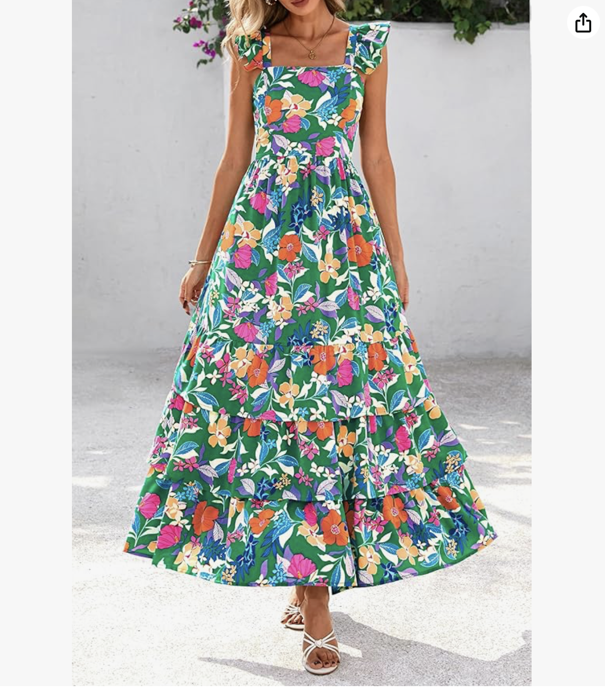 Woman wearing a long, colorful floral dress with ruffled sleeves, standing outdoors on a concrete surface with a plain wall and some purple flowers in the background.