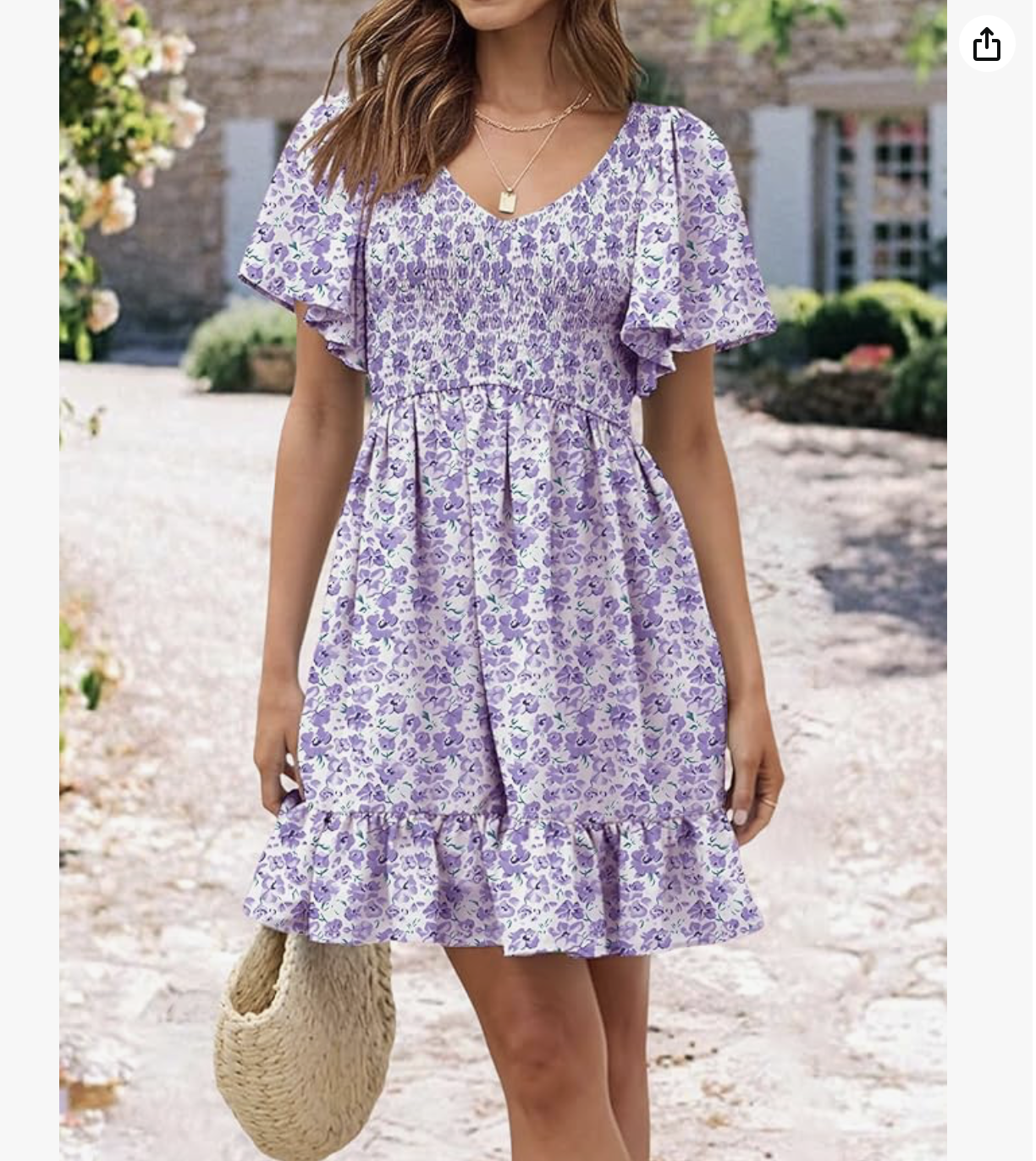 A woman wearing a purple floral dress with ruffled sleeves and hem, holding a woven straw bag, standing outdoors in a garden with a gravel path and green plants.