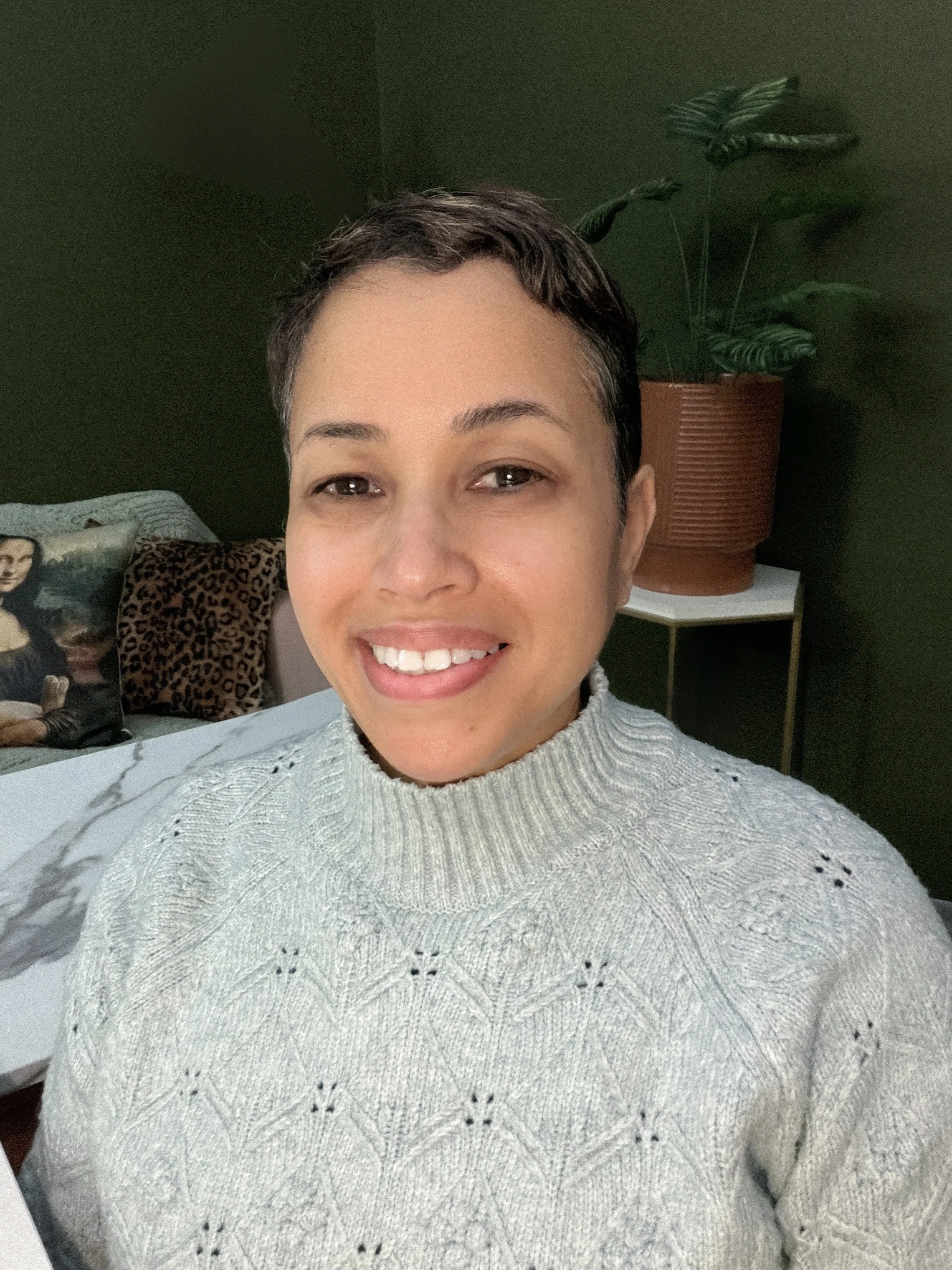A woman with short dark hair smiling indoors, wearing a light gray knitted sweater, with a dark green wall, a potted plant, a bed with pillows, and a table in the background.
