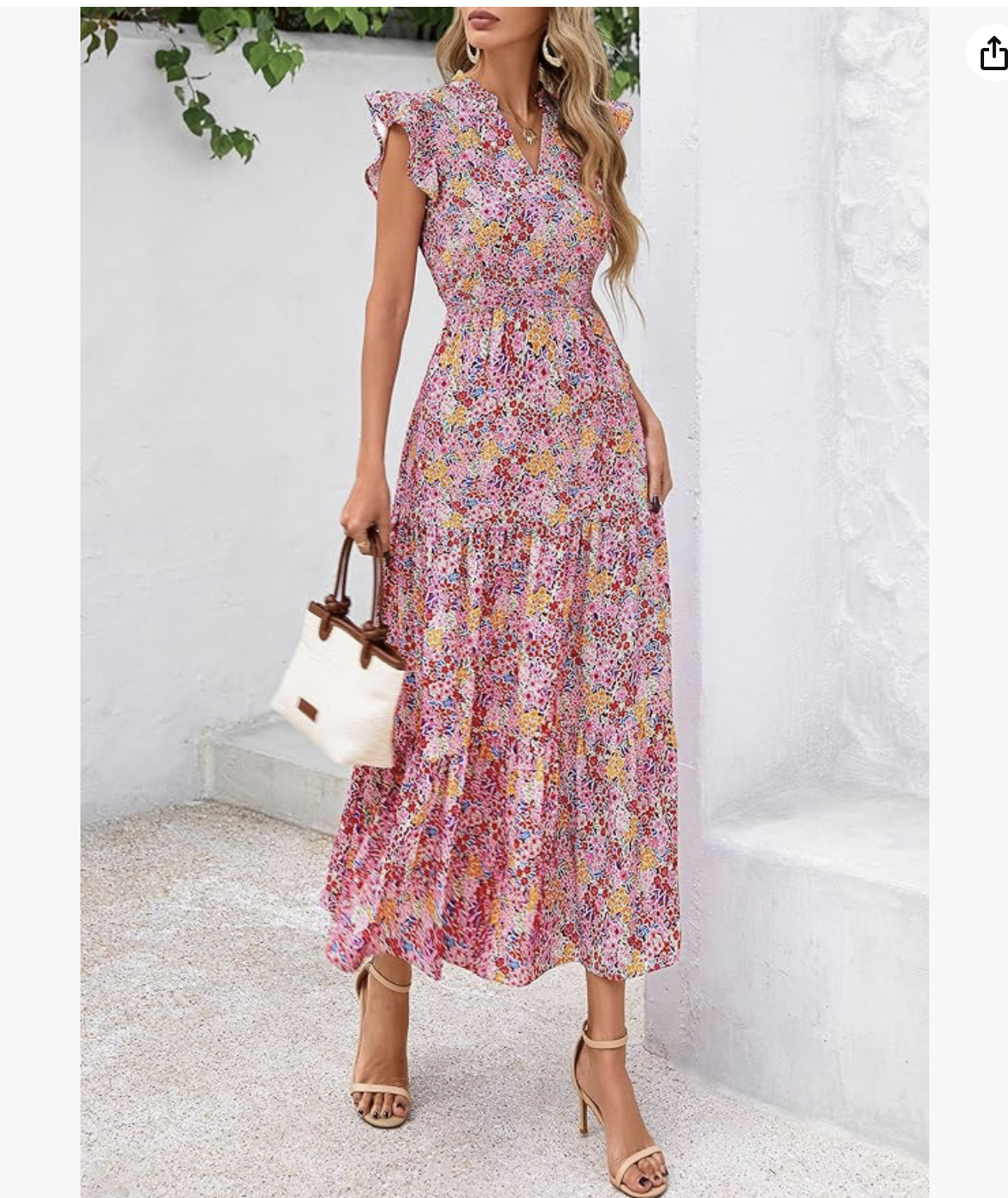 A woman in a colorful floral maxi dress holding a white handbag with wooden handles, standing against a white textured wall and greenery.