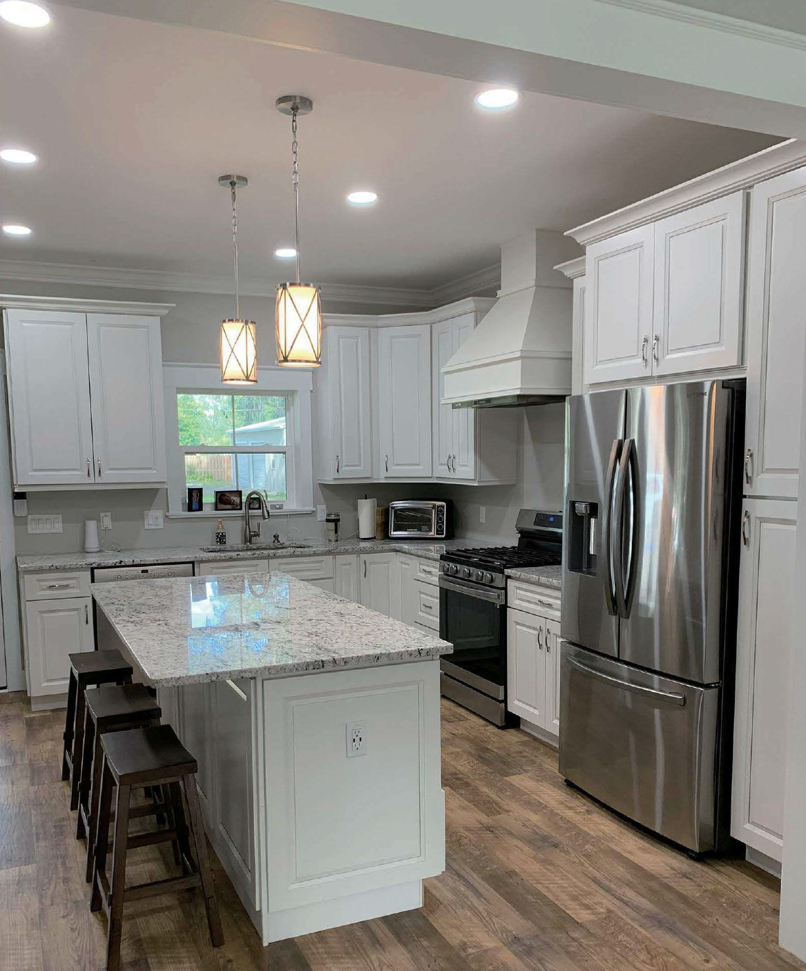 Modern kitchen with white cabinets, stainless steel refrigerator, oven, microwave, and island with three dark stools. Pendant lights hang over the island, with recessed ceiling lights, and a window above the sink looking outside.