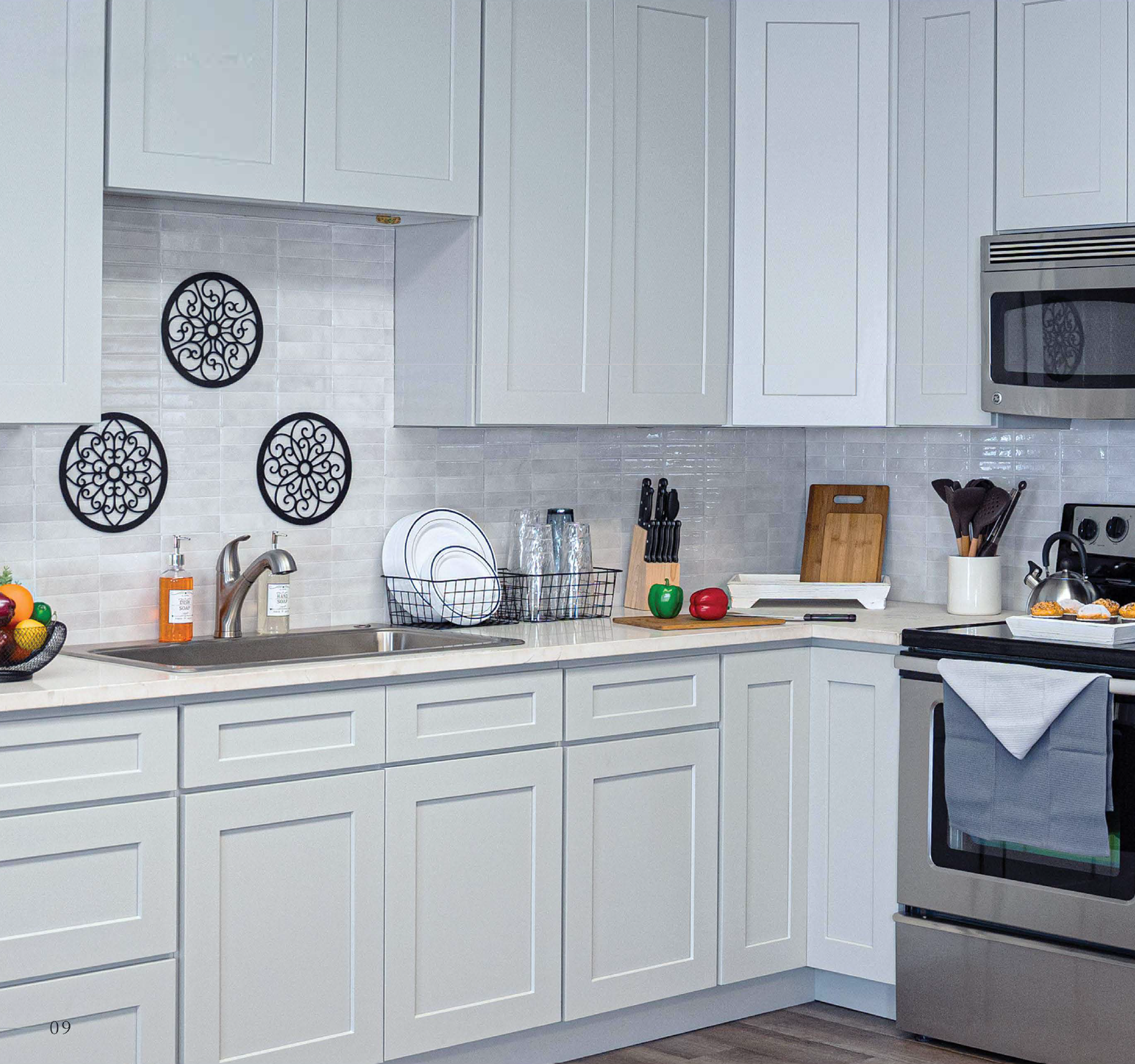 White kitchen with white cabinets, a stainless steel stove, microwave, and a single sink. Countertop holds plates, glasses, cutting board, apples, and a basket of fruit. Decor includes black wall art and a white backsplash.