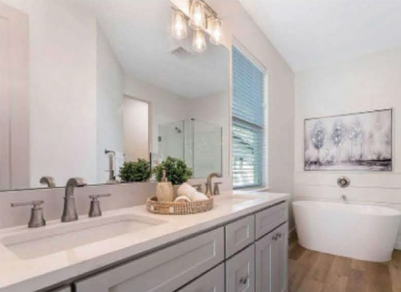 Bathroom with a double sink vanity, large mirror, potted plants, soap dispenser, window with blinds, and a standalone bathtub.