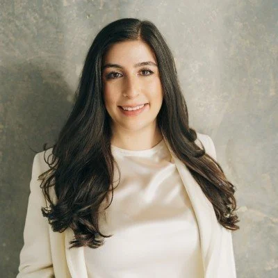 A young woman with long dark hair wearing a white blazer and smiling, standing against a gray textured background.