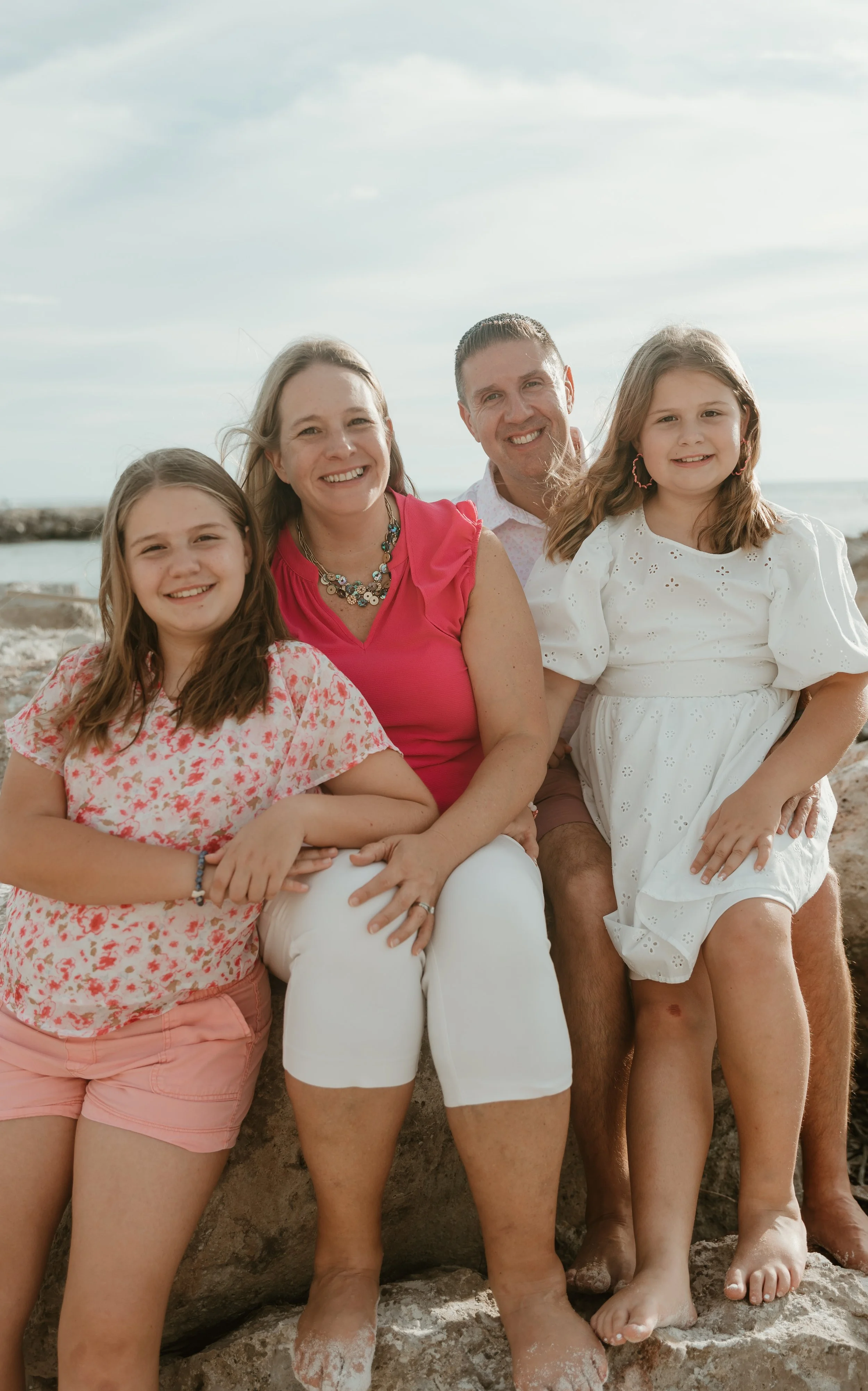 A family of four at the beach, sitting on rocks, smiling for a photo with the ocean and sky in the background.