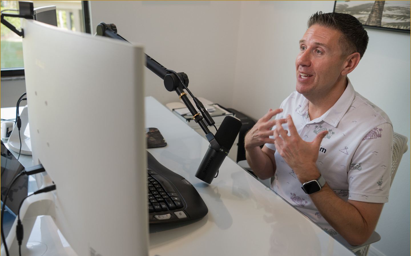A man wearing a white shirt with a nautical pattern, sitting at a desk, speaking into a microphone, and gesturing with his hands. He is in a bright office with a window and a framed picture on the wall.