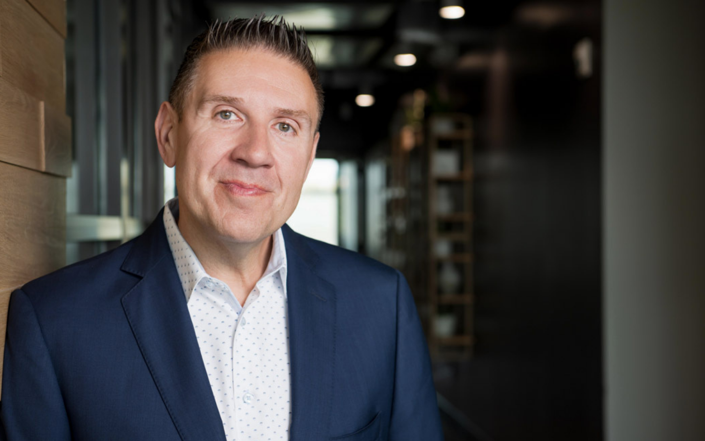 A middle-aged man in a navy blazer and white shirt with small polka dots, standing indoors near a wooden wall, with a blurred background of modern interior decor.