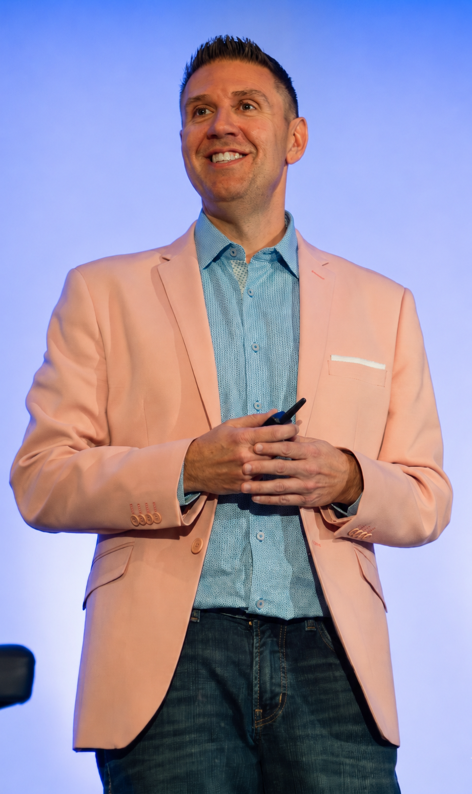 A man smiling while holding a remote, wearing a light pink blazer over a blue patterned shirt, standing against a blue background.