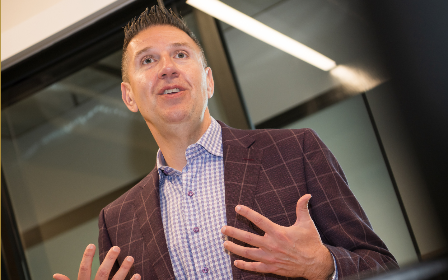 A man in a checked dress shirt and plaid blazer speaking, gesturing with his hands, in an indoor setting.