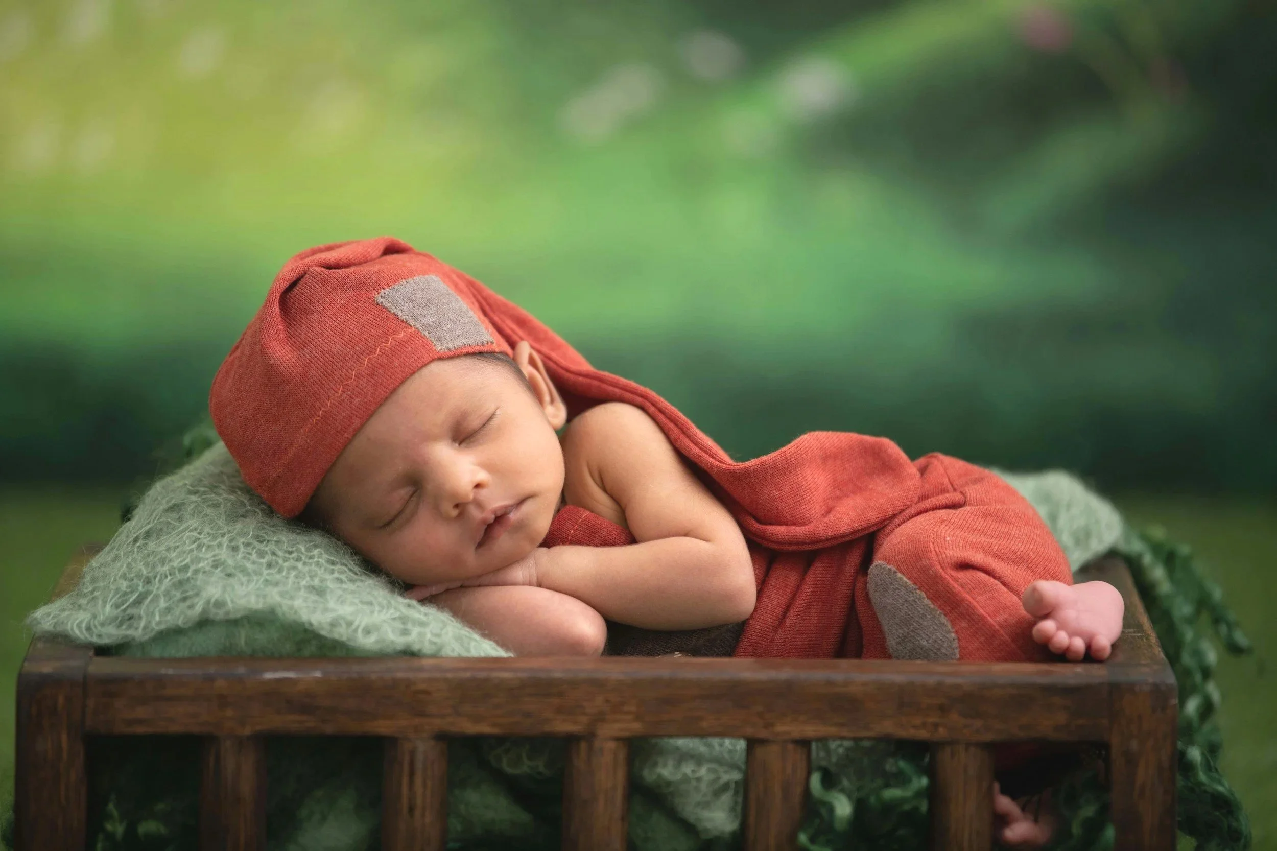 A sleeping baby boy wearing a red hat and matching red outfit, laying on a green blanket in a small wooden bed with a blurred outdoor background.