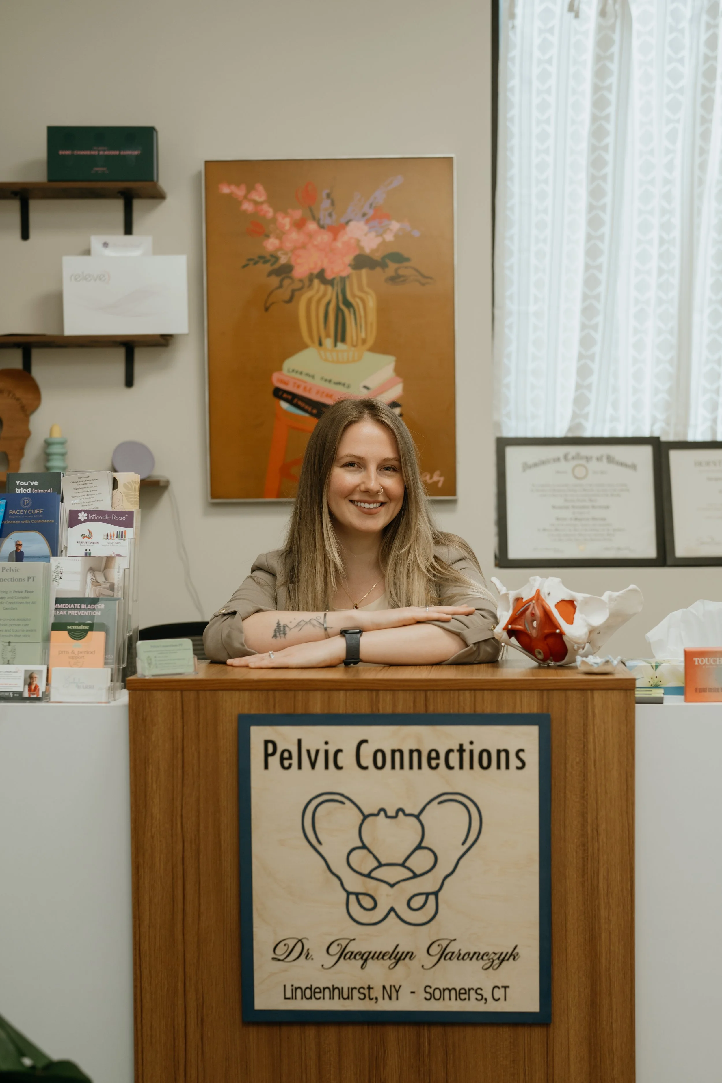 Reception desk for Pelvic Connections with a smiling woman sitting behind it. The desk has a sign with the clinic's name and a pelvic diagram. There are medical brochures on the desk, and the background features wall art with a floral vase and books, along with framed certificates and a window with patterned curtains.