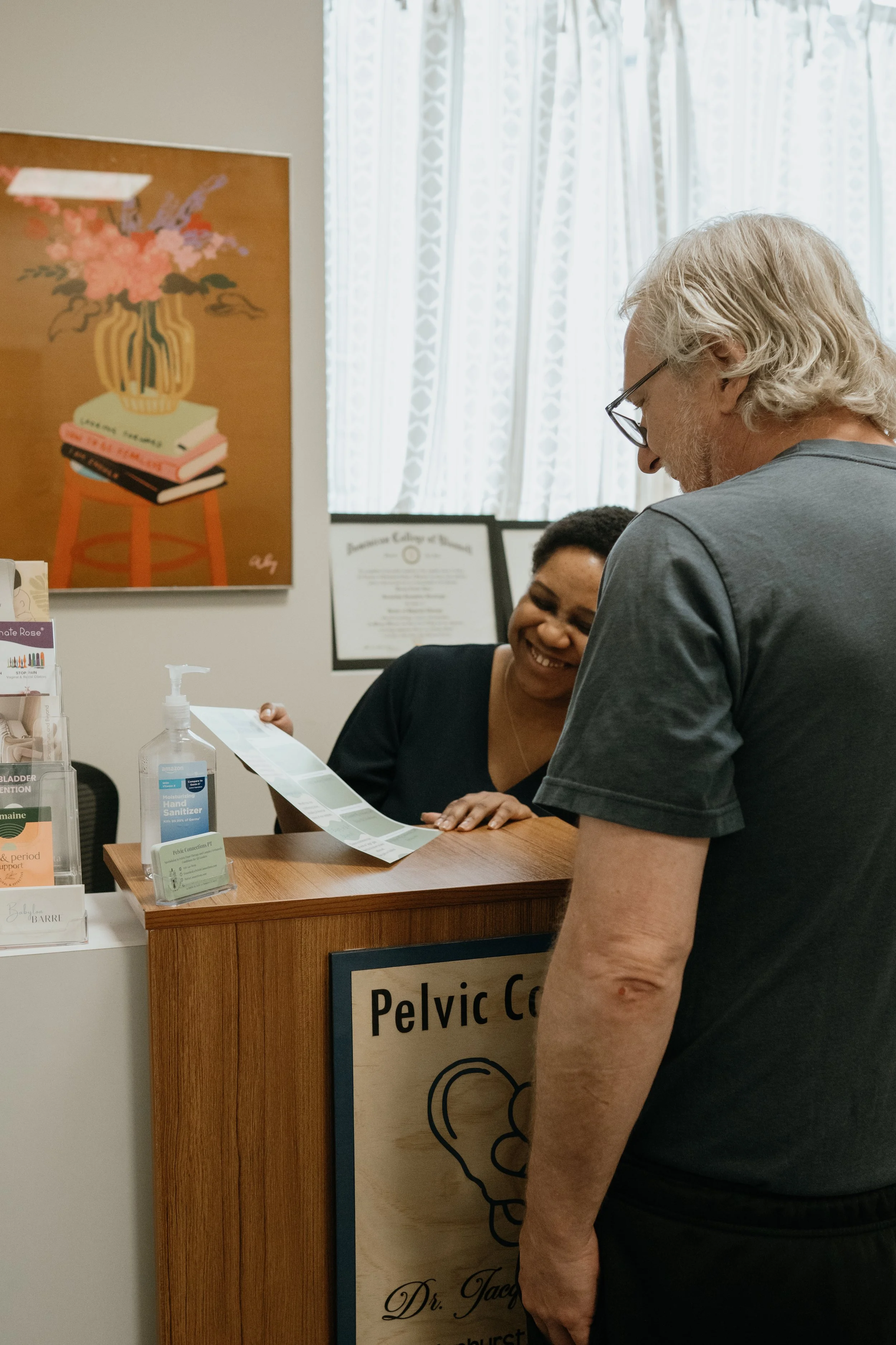 A man with long gray hair and glasses looking at a woman behind a reception desk in a medical office. The woman is smiling and handing him a paper. The desk has hand sanitizer, brochures, and a sign that partially reads "Pelvic.