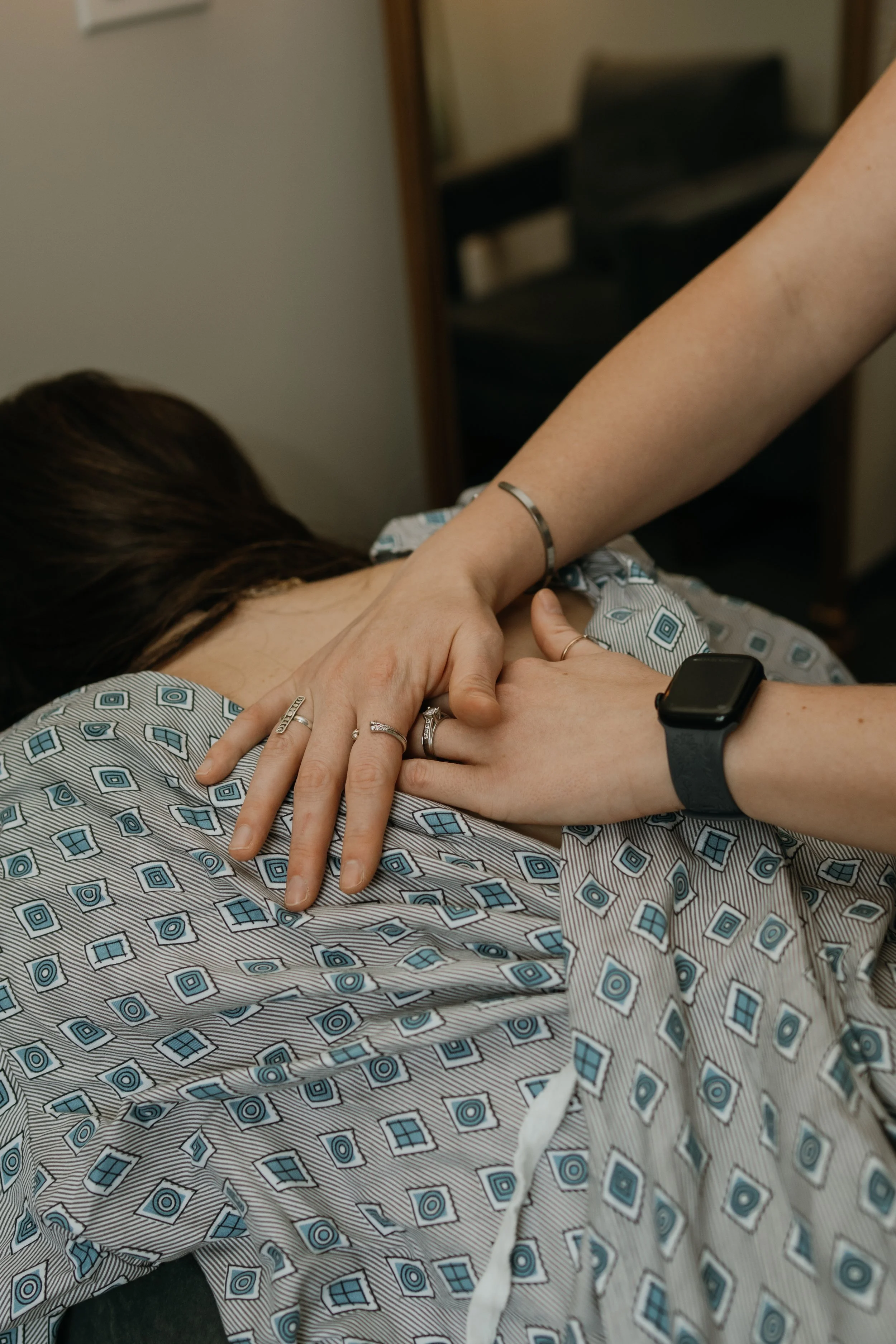 A person lying in a hospital bed with a patterned hospital gown, holding hands with another person, showing rings on their fingers. The person in bed has dark hair, and the arm of the person holding hands has a smartwatch on the wrist.