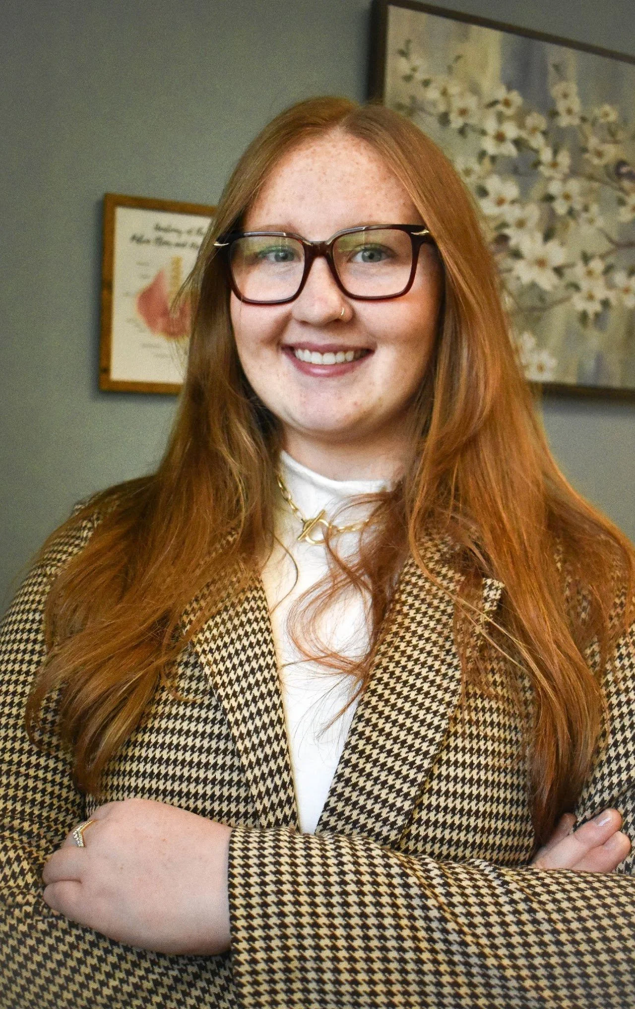 A young woman with long red hair, wearing glasses, a white turtleneck, a houndstooth blazer, gold jewelry, and smiling with arms crossed in front of a wall with framed pictures.