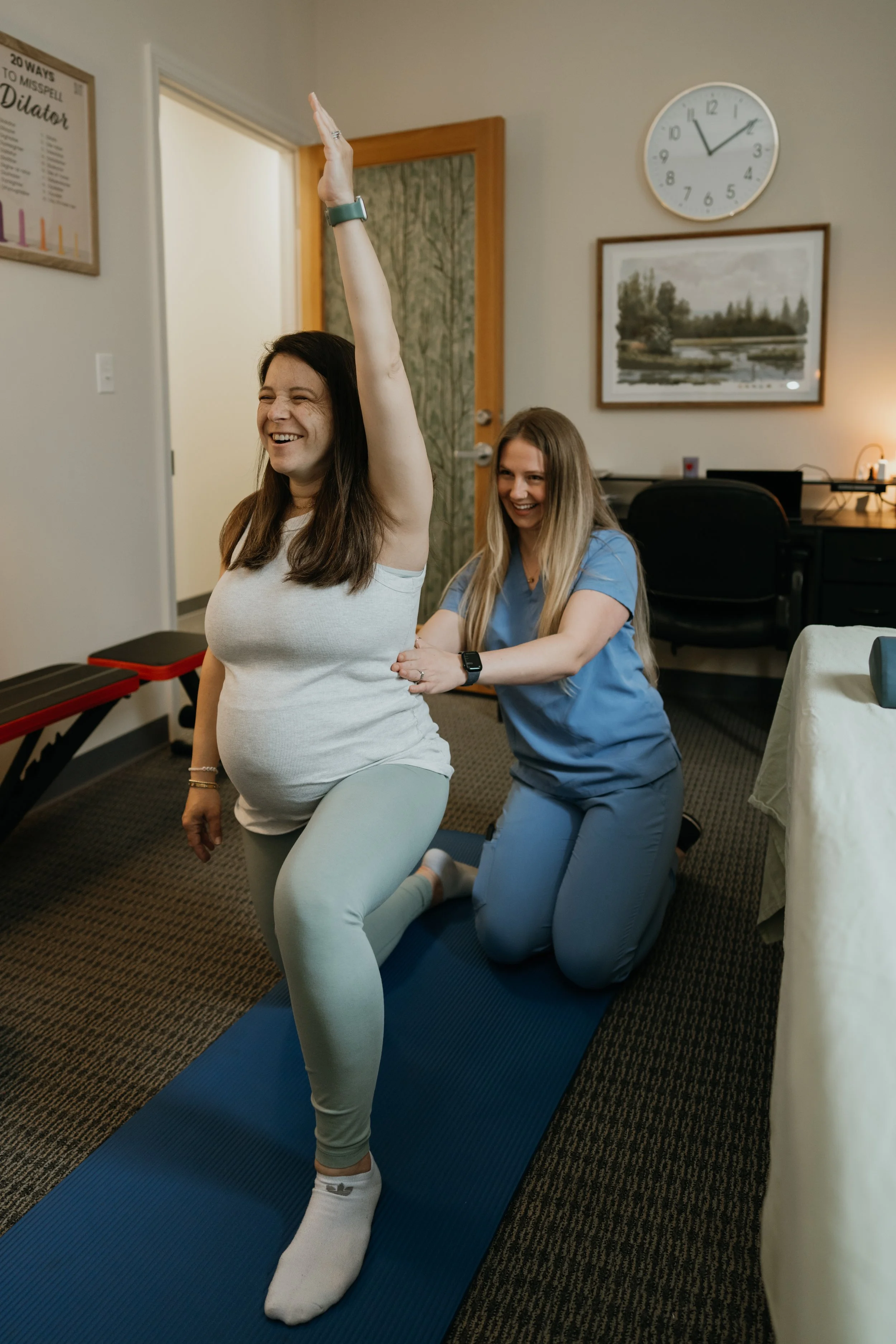 Pregnant woman performing a yoga pose on a blue mat while a healthcare professional assists her in an examination room.
