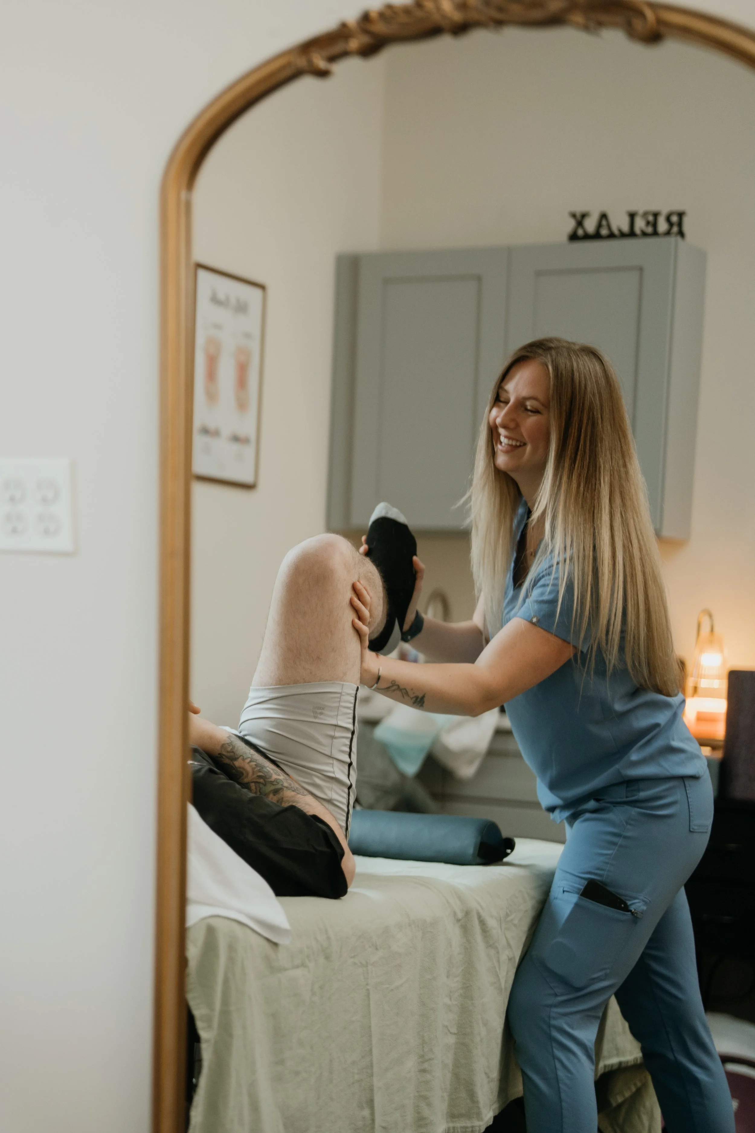 A medical professional in blue scrubs caring for a patient lying on a bed, as seen through a mirror.