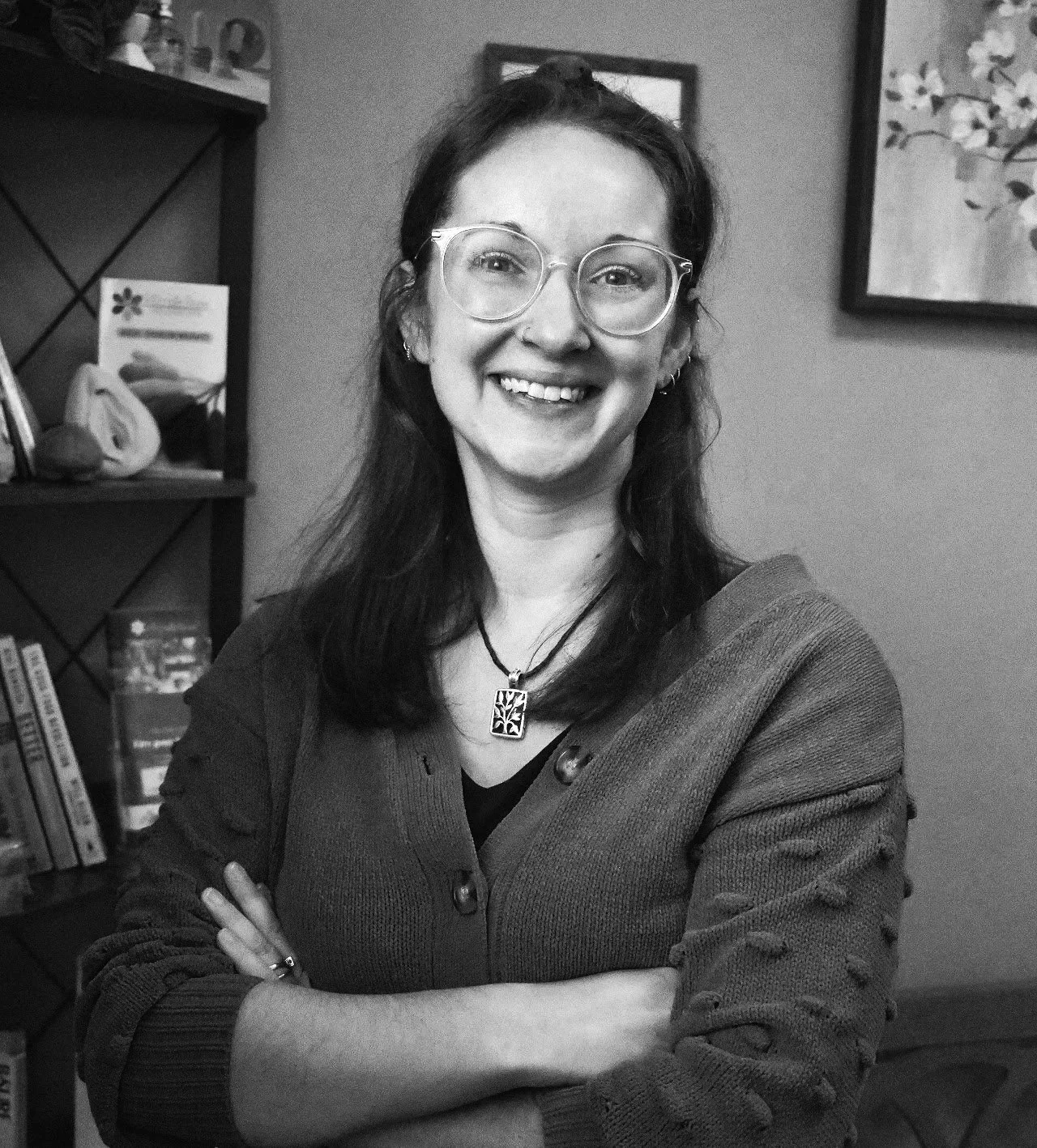 A woman with glasses and long dark hair smiling, arms crossed, standing in a room with bookshelves and framed pictures in the background.