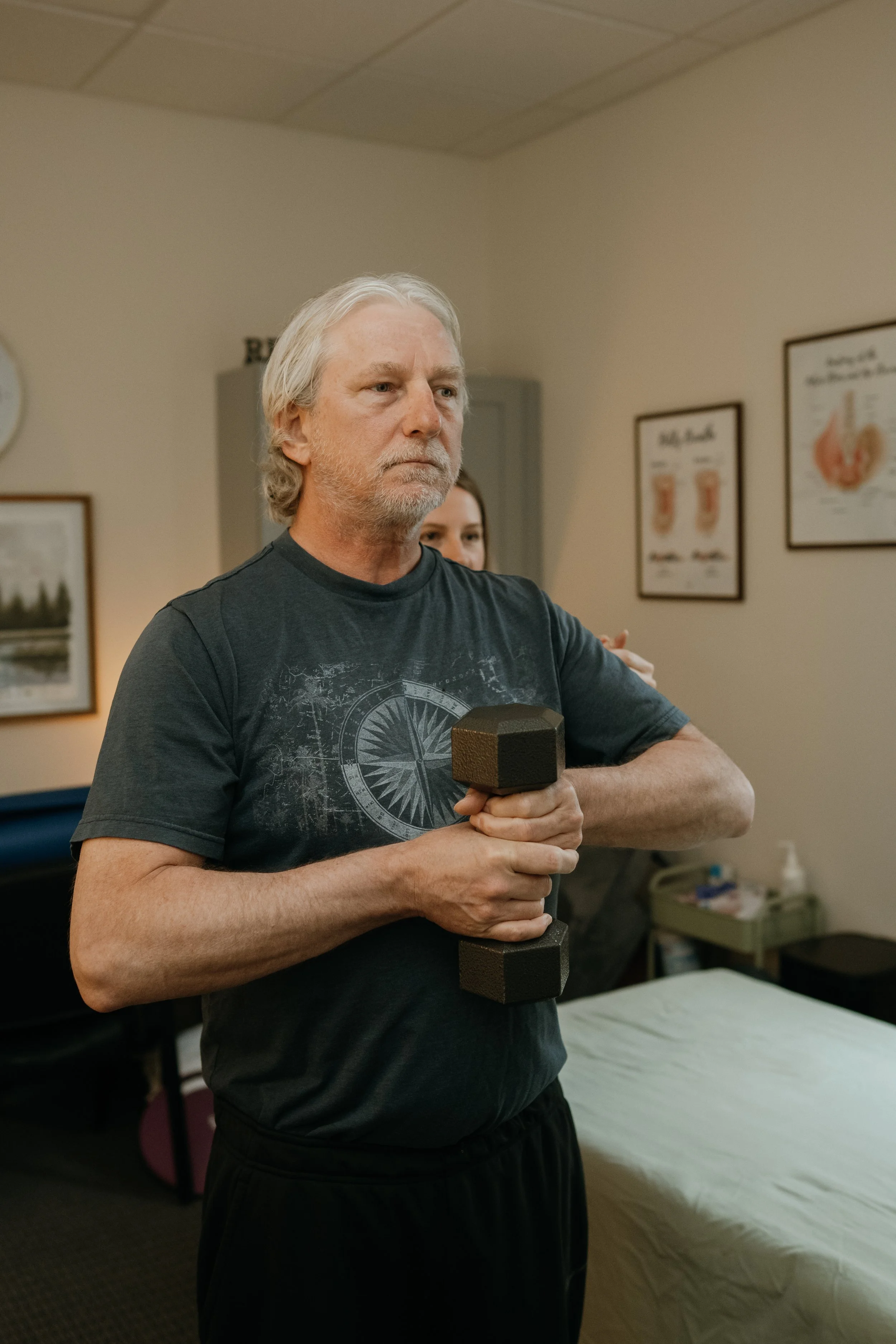 An older man with gray hair and a beard holding a dumbbell in a room with beige walls, medical posters, and a bed with a cream-colored bedspread.