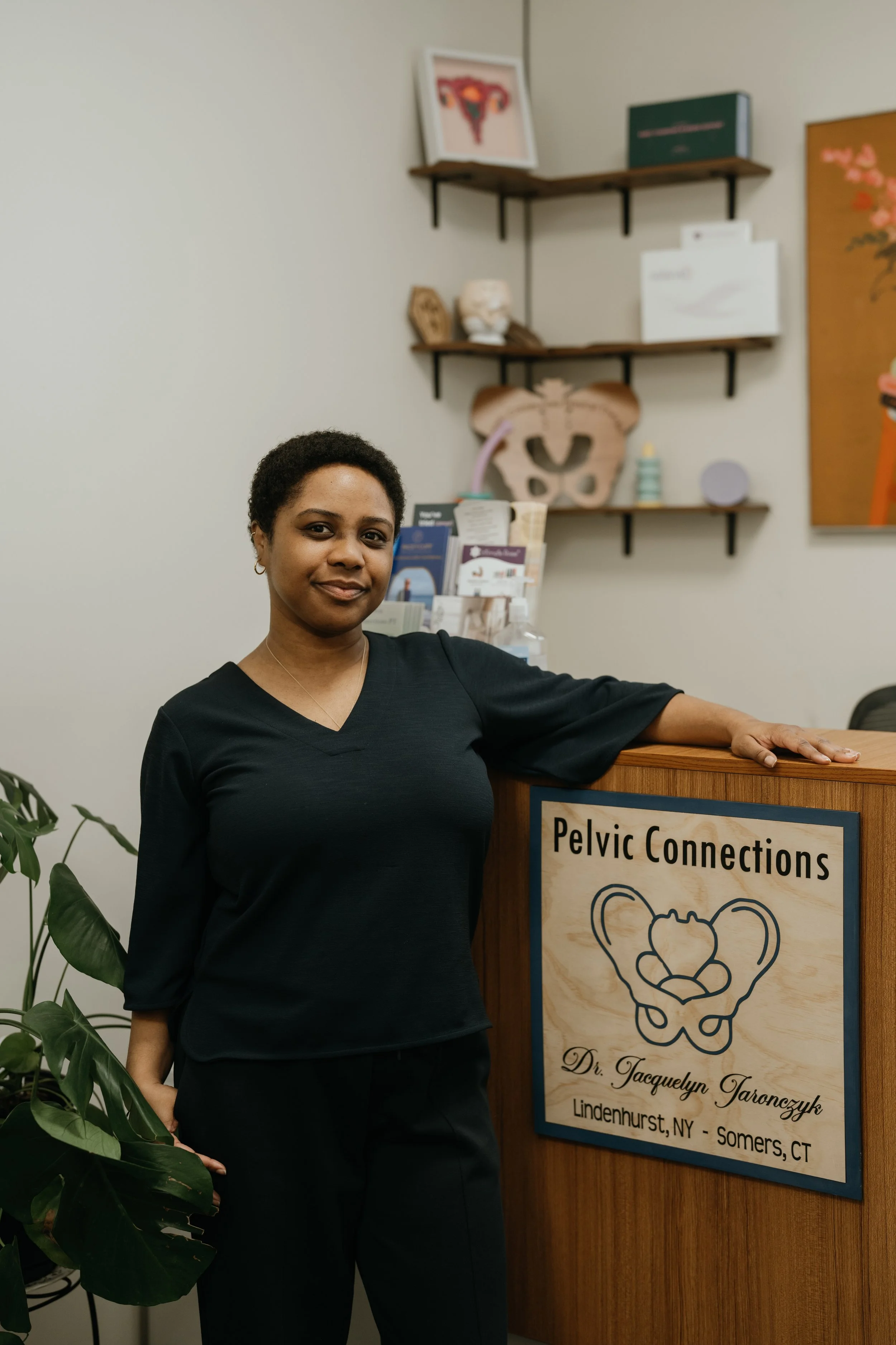 A woman with short curly hair wearing a black top, standing in front of a wooden reception desk with a sign that reads "Pelvic Connections" and an illustration of a pelvis. The woman is smiling slightly and resting her arm on the desk. Behind her are wall shelves with medical models, books, and decorative items, and part of a framed art piece is visible on the wall.