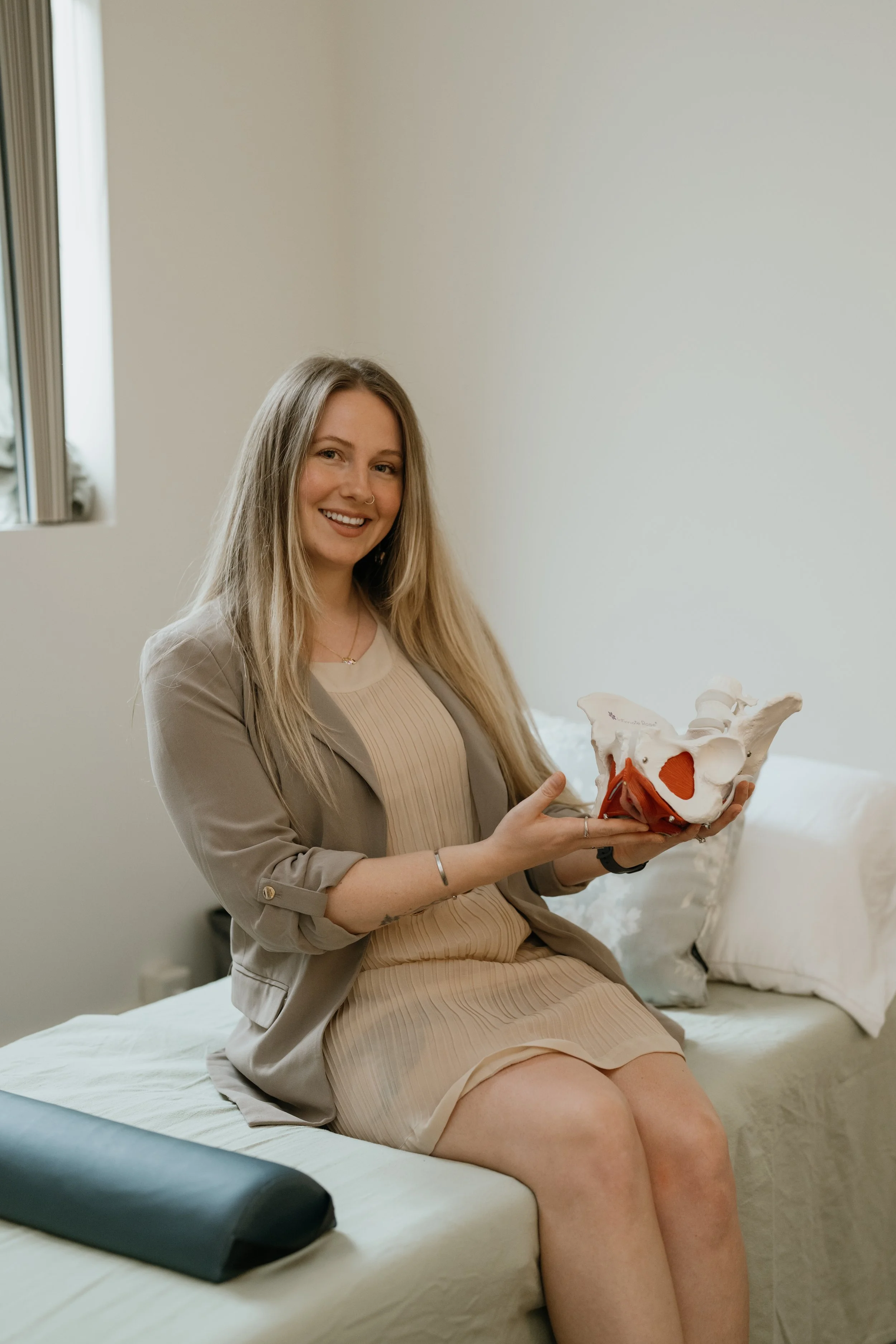 A woman sitting on a bed in a beige dress and blazer holding a pelvic model, smiling at the camera.