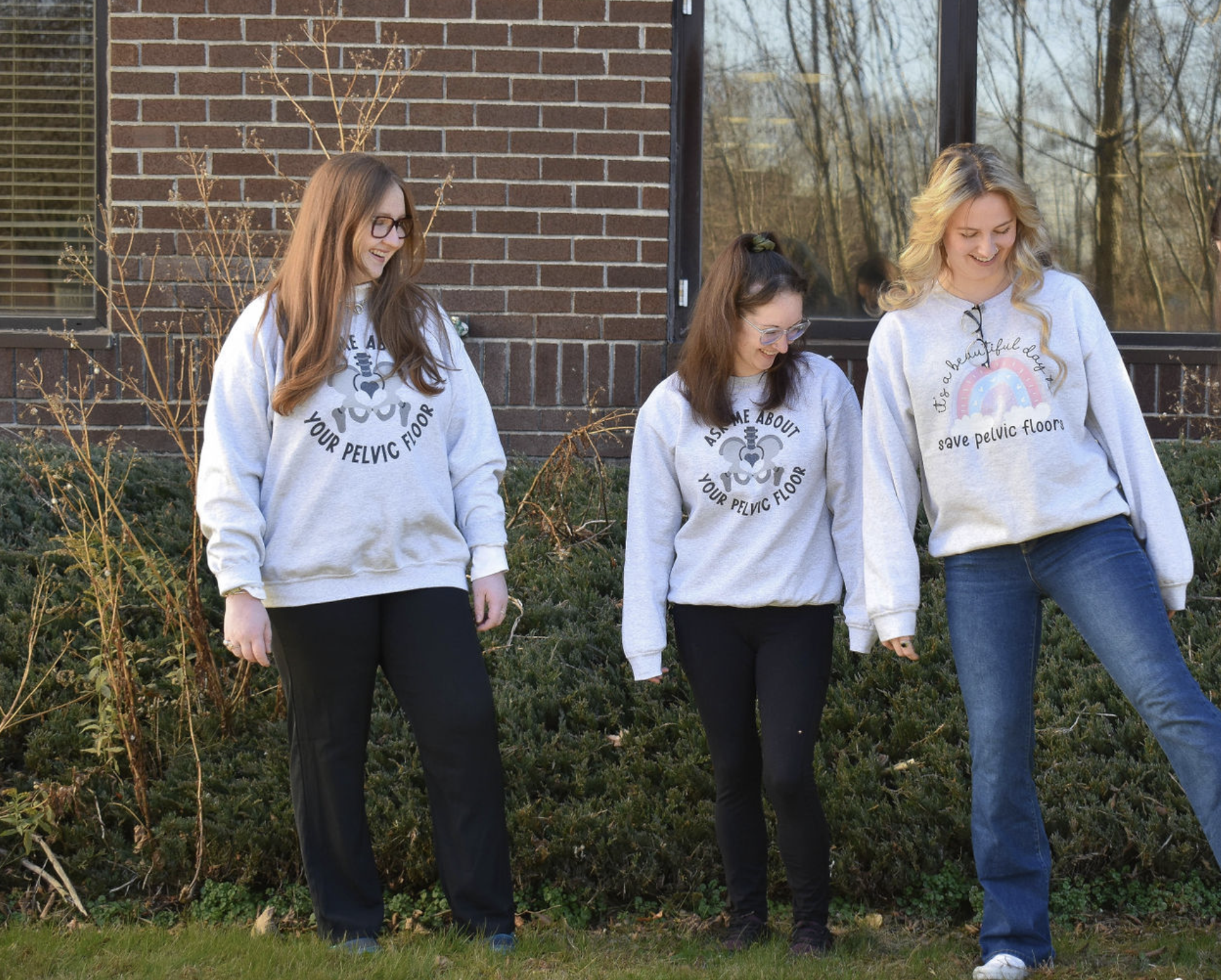 Three women walking outdoors near a brick building during autumn, wearing gray sweatshirts with a band and text about pelvic health, smiling and looking down.