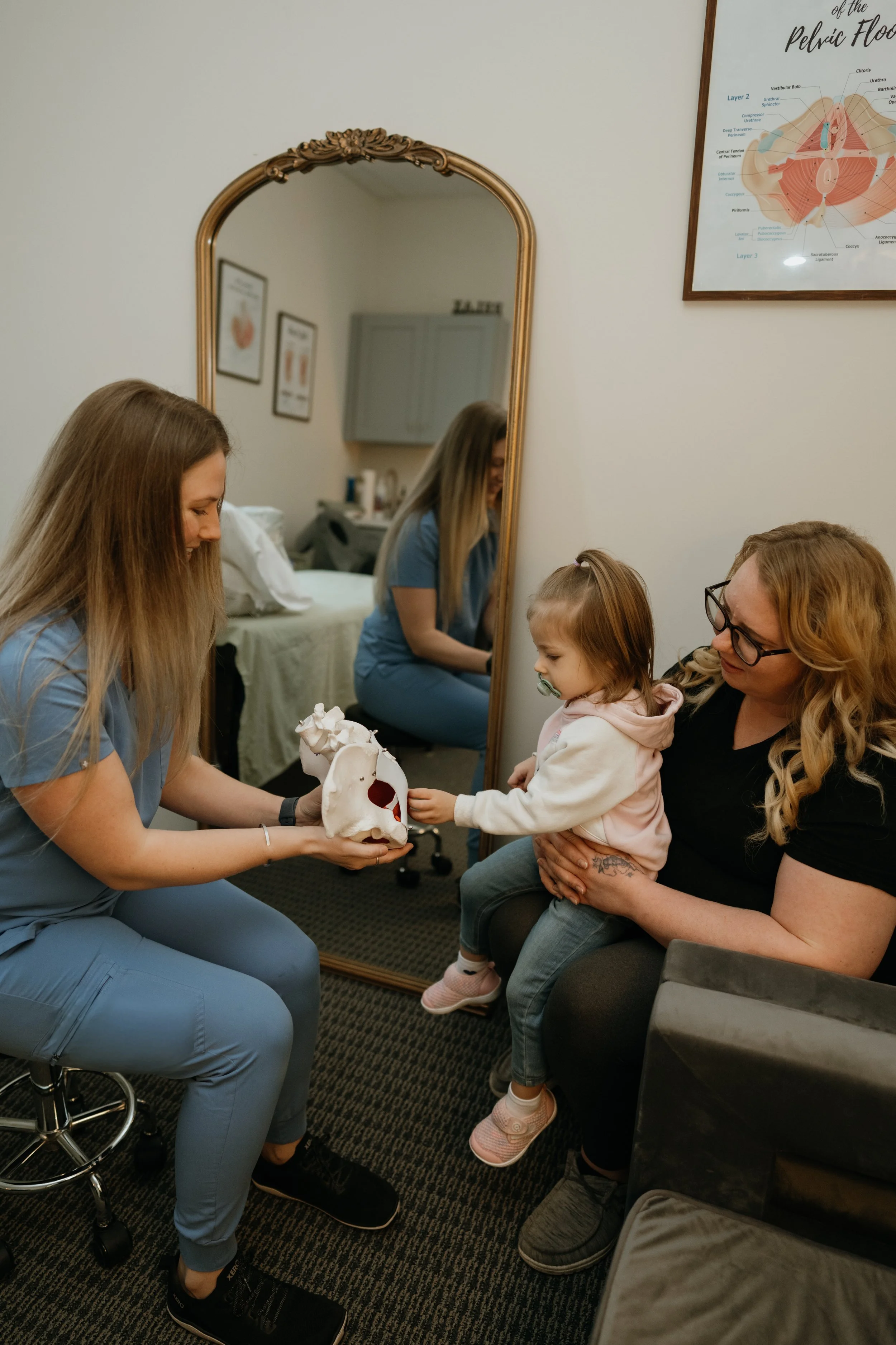 A healthcare professional holding a pelvic bone model while sitting on a stool, engaging with a young girl and a woman in a medical examination room. A mirror reflects the scene, and a pelvic anatomy poster hangs on the wall.