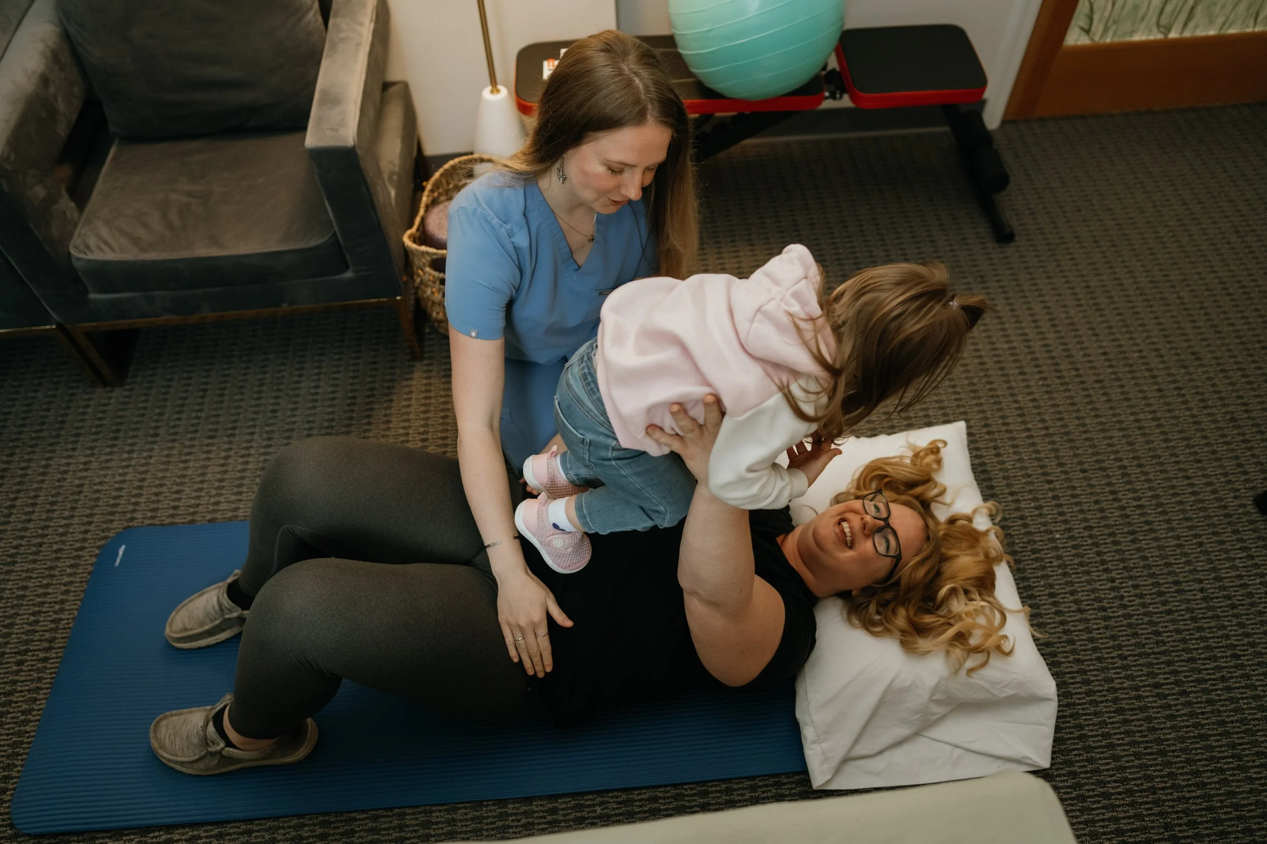 A woman lying on a black massage table with two young children, one on her chest and the other on her legs, in a room with a couch and exercise equipment.