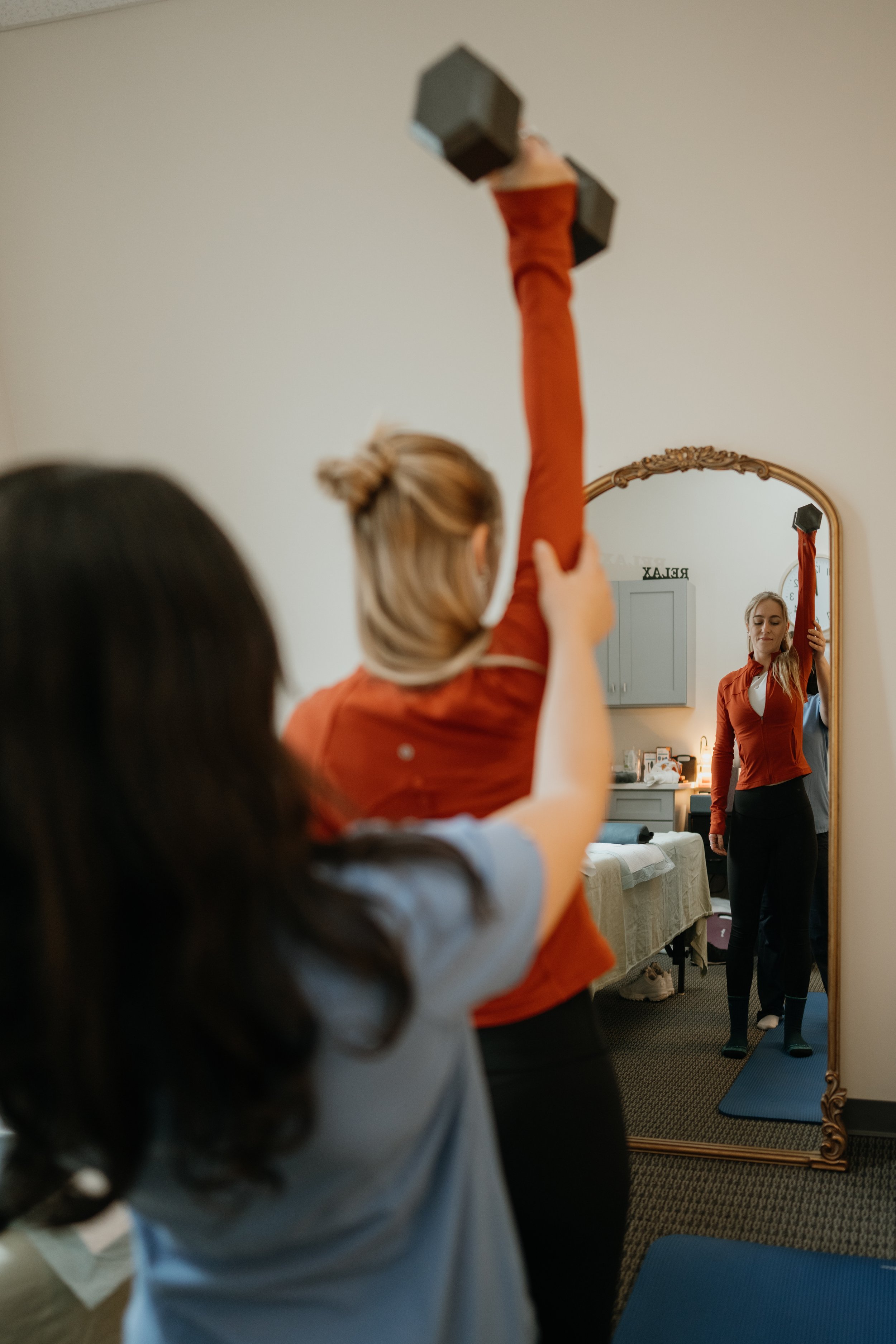 A woman in a rust-colored jacket lifting a dumbbell in front of a mirror, with a second person's arm visible, in a room with a dresser, mirror, and blue exercise mat.