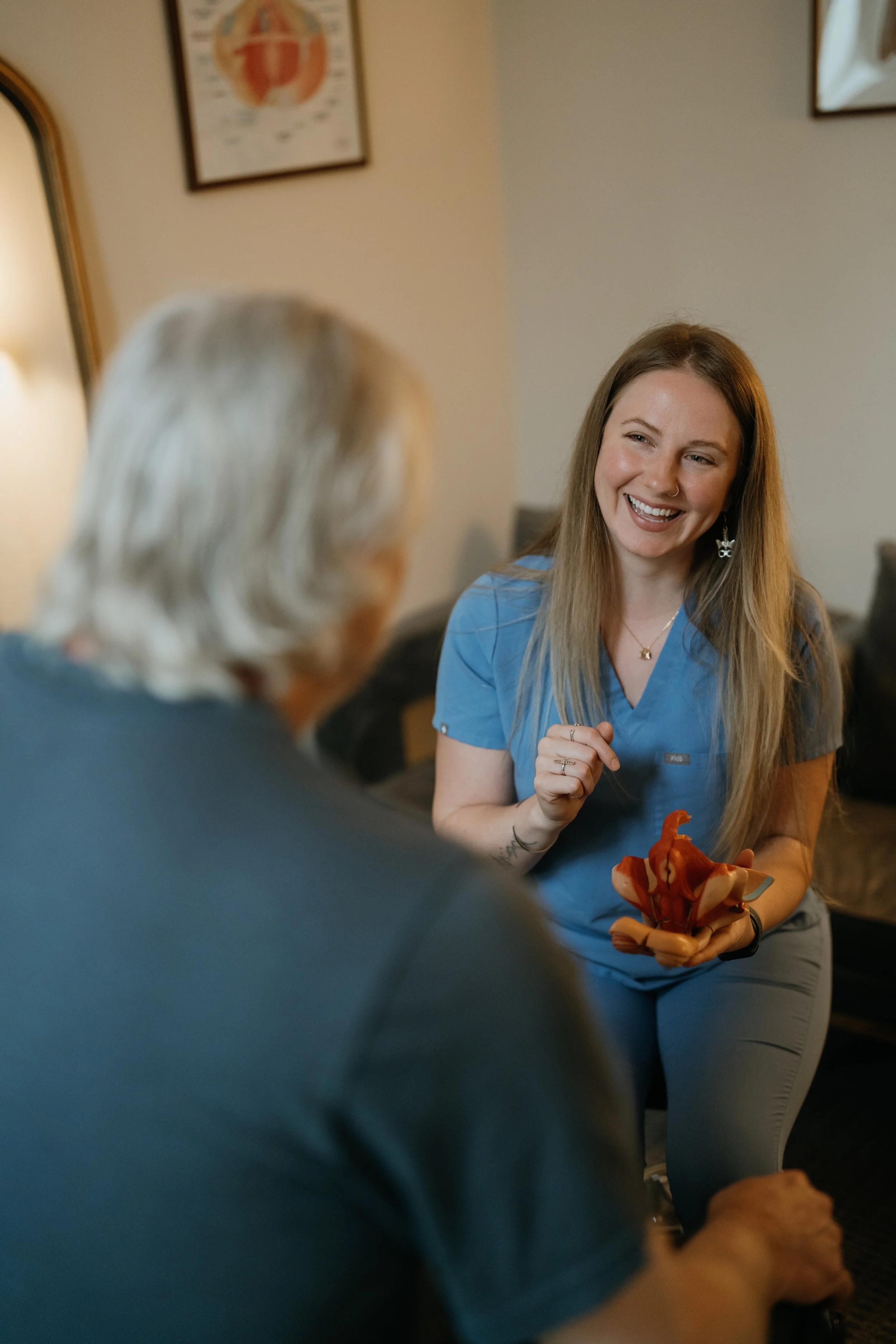 A young woman in medical scrubs smiling and talking with an older person in a room with framed pictures on the wall.