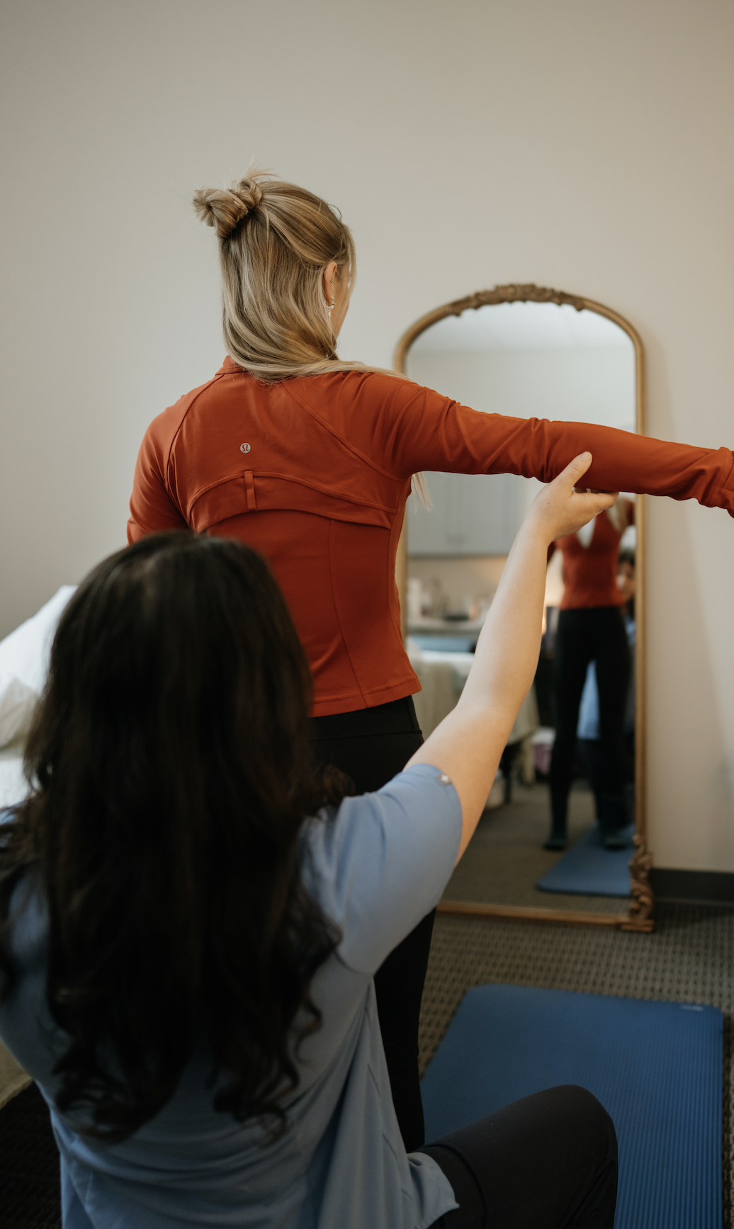 A woman in a red jacket is doing a stretching exercise in front of a mirror, guided by a woman sitting on a yoga mat in a room.