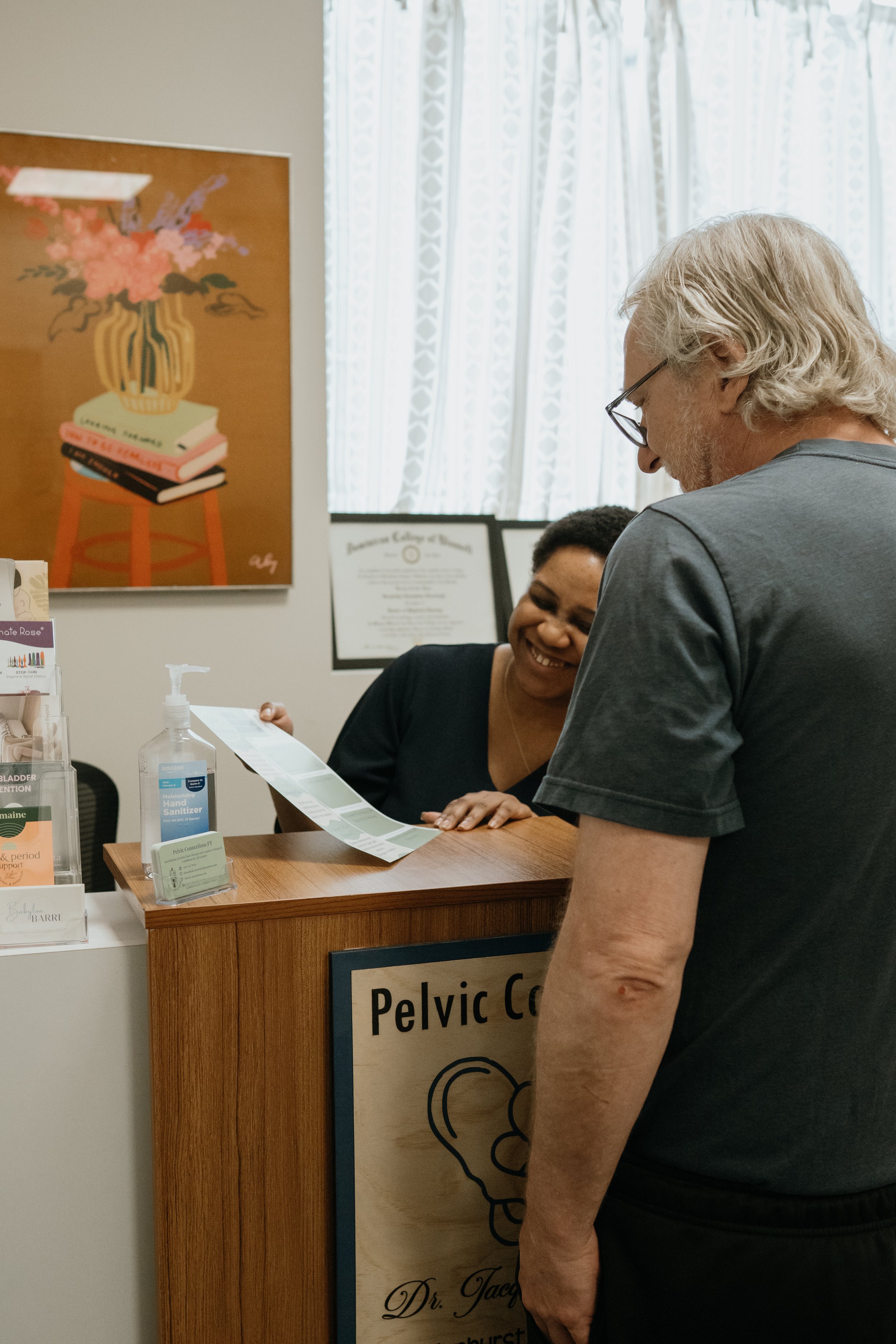 A man with gray, curly hair wearing glasses and a dark t-shirt talking to a woman behind a reception desk at a medical office, with a sign reading 'Pelvic Co' and a poster of a brain and spine on the wall.