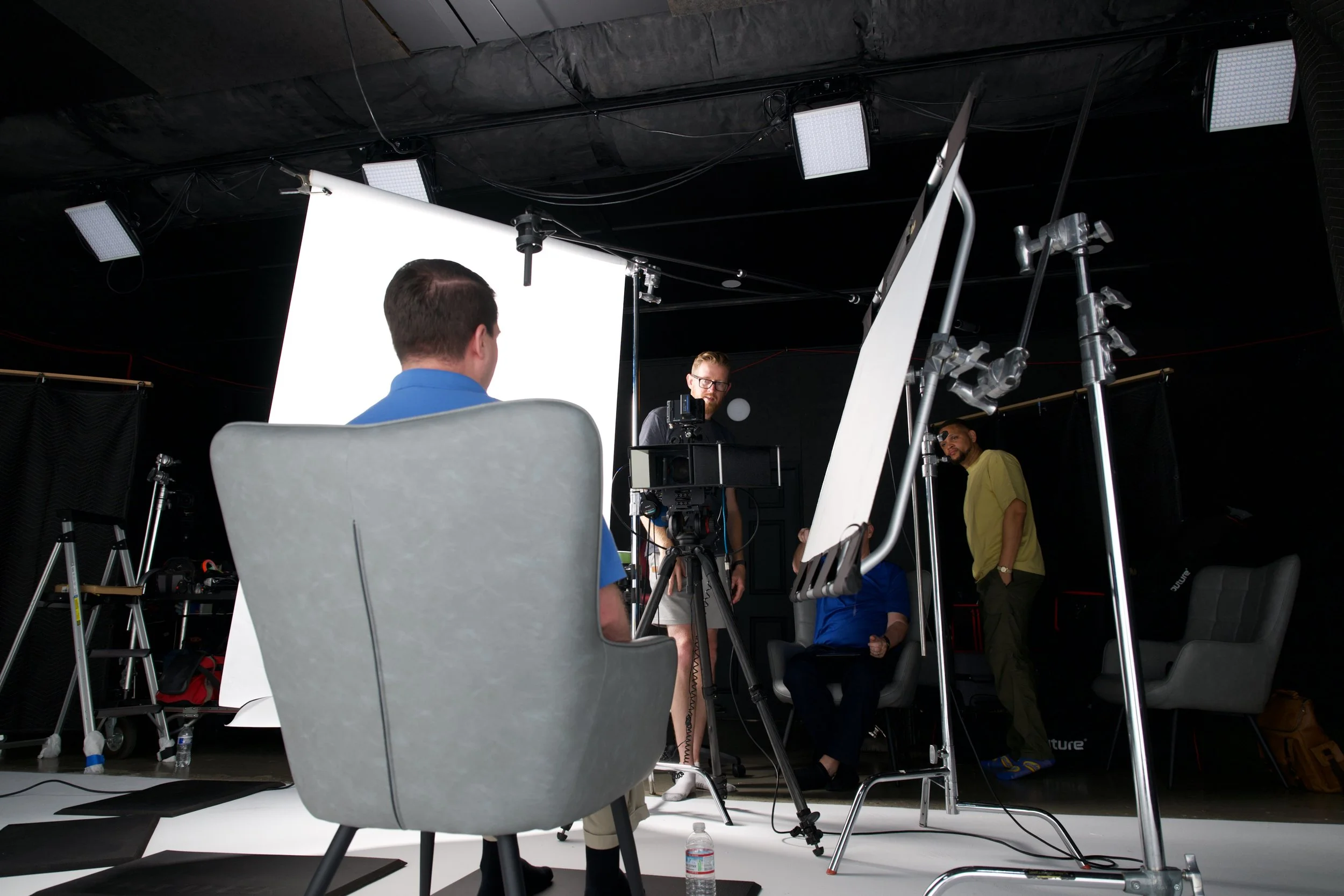 People conducting an interview in a professional studio setup with lighting and equipment, one person seated in front of the camera and two others standing or sitting nearby.