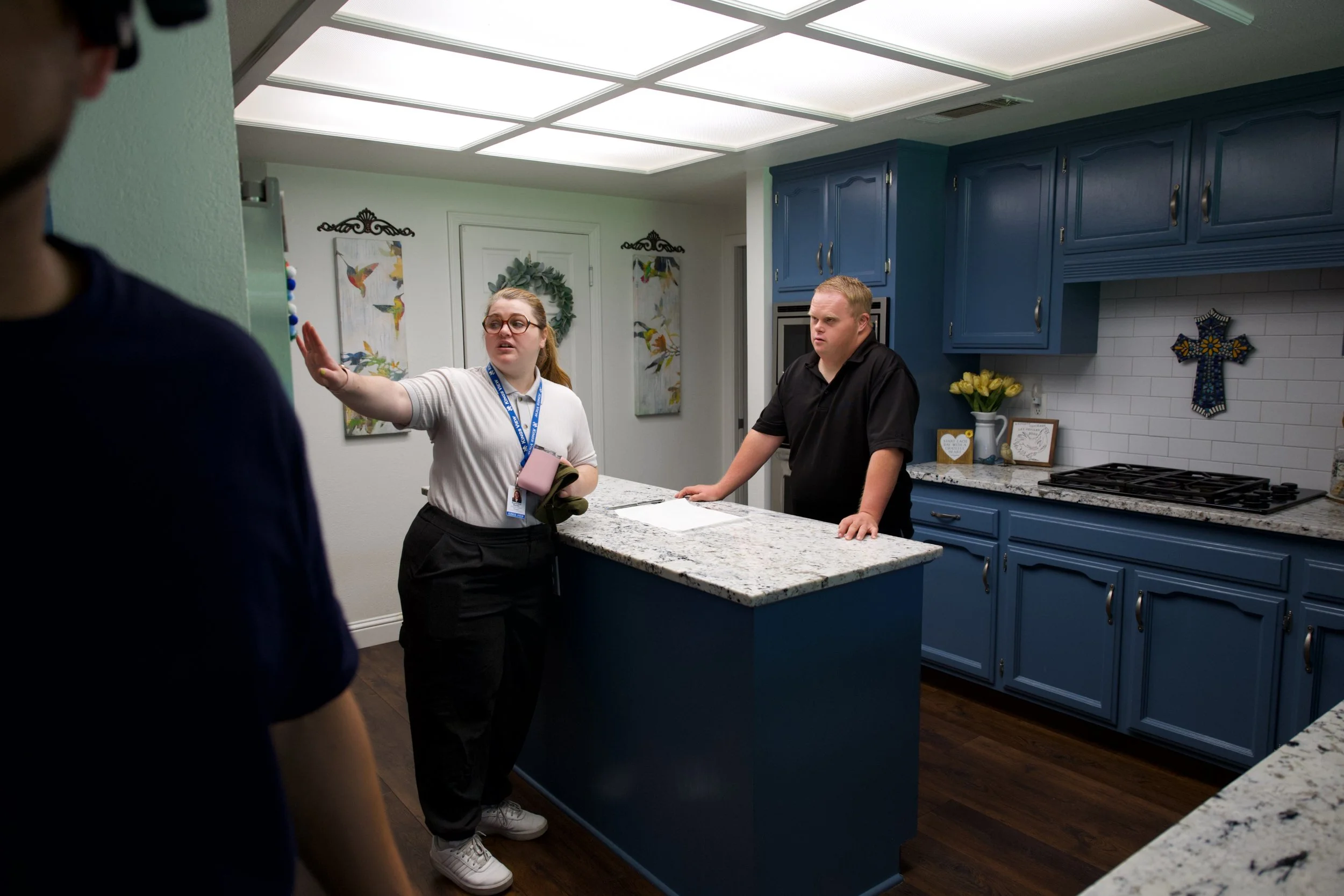 A woman with glasses and a lanyard is speaking and gesturing with her hand to a man standing behind a kitchen counter. The man is leaning on the counter with his hands and looks attentive. Another person in a dark shirt is partially visible in the fo