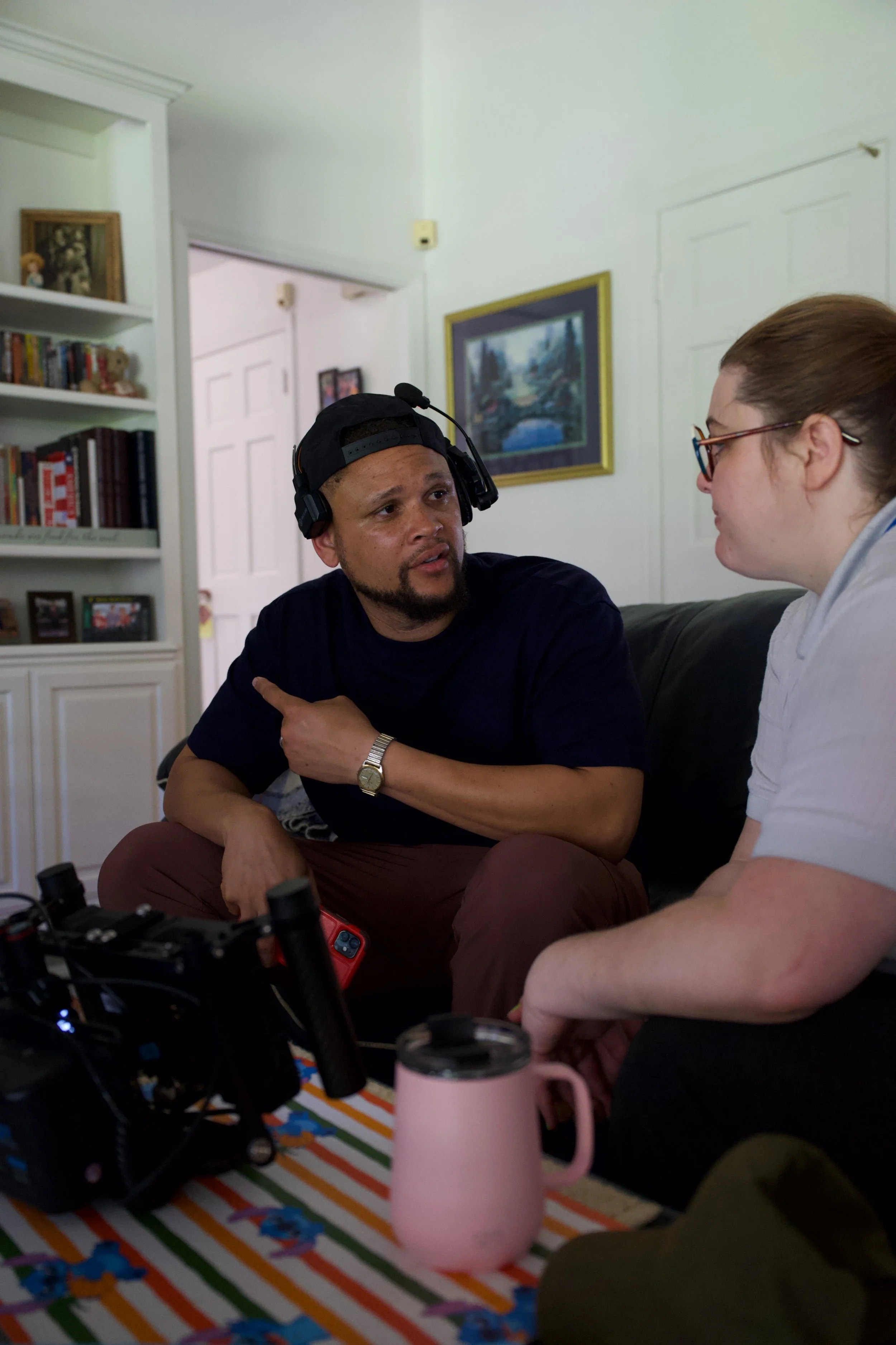 Two people having a conversation in a living room, one with a headset, sitting on a couch, and the other with glasses and a white shirt. A table with a pink mug and camera equipment in front of them.