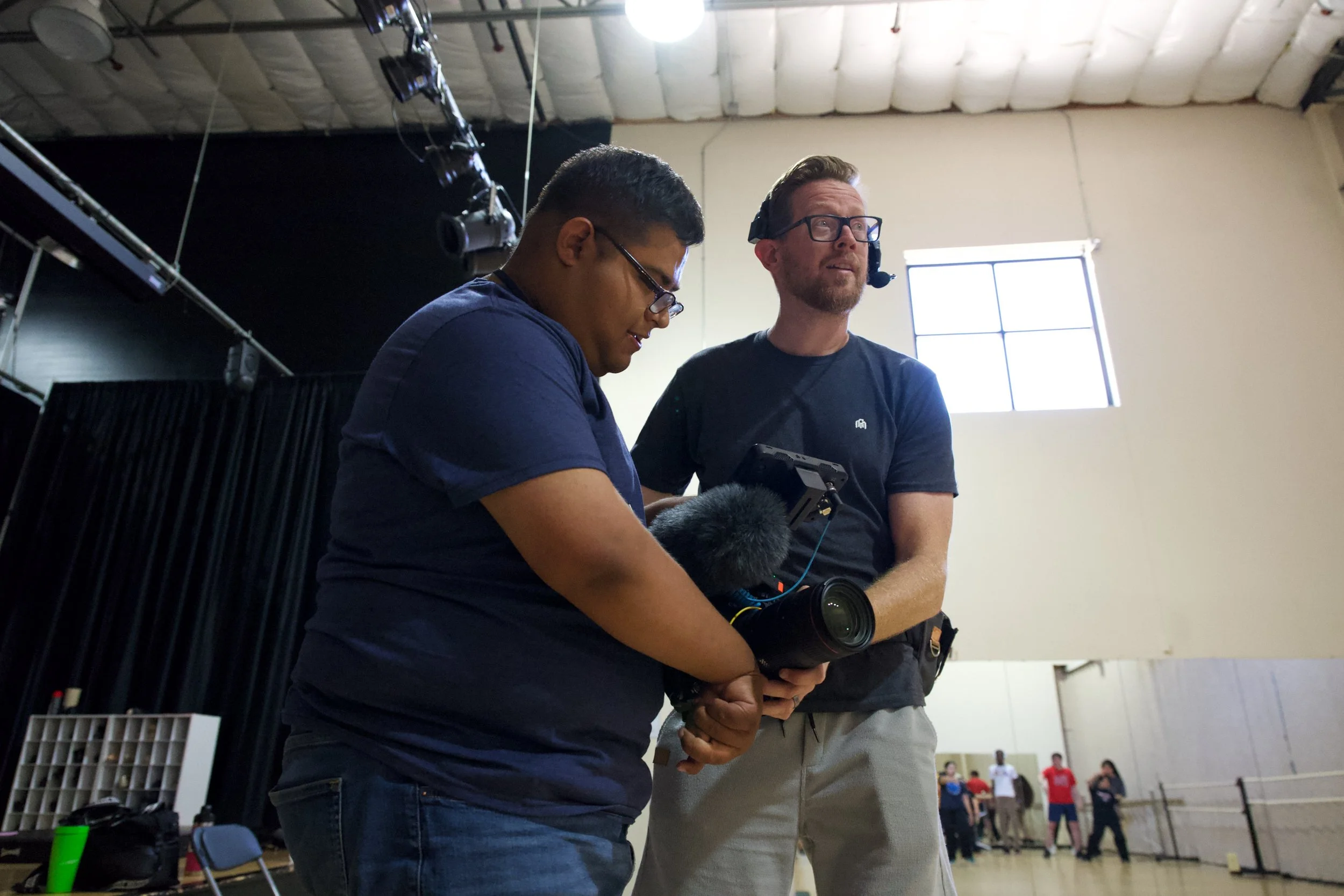 Two men working together on video filming equipment inside a room with a high ceiling, black curtains, and a window.