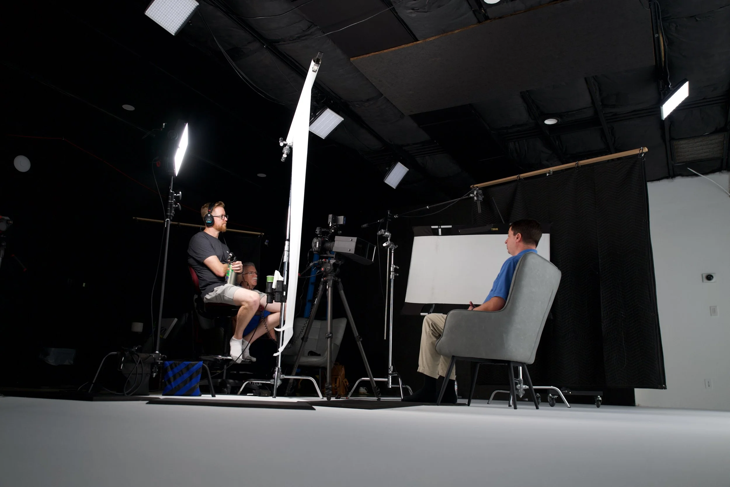 People filming an interview or recording in a studio with professional lighting and equipment. One person is seated in a gray armchair, and two others are seated nearby, one with headphones and a beverage, engaging in conversation.