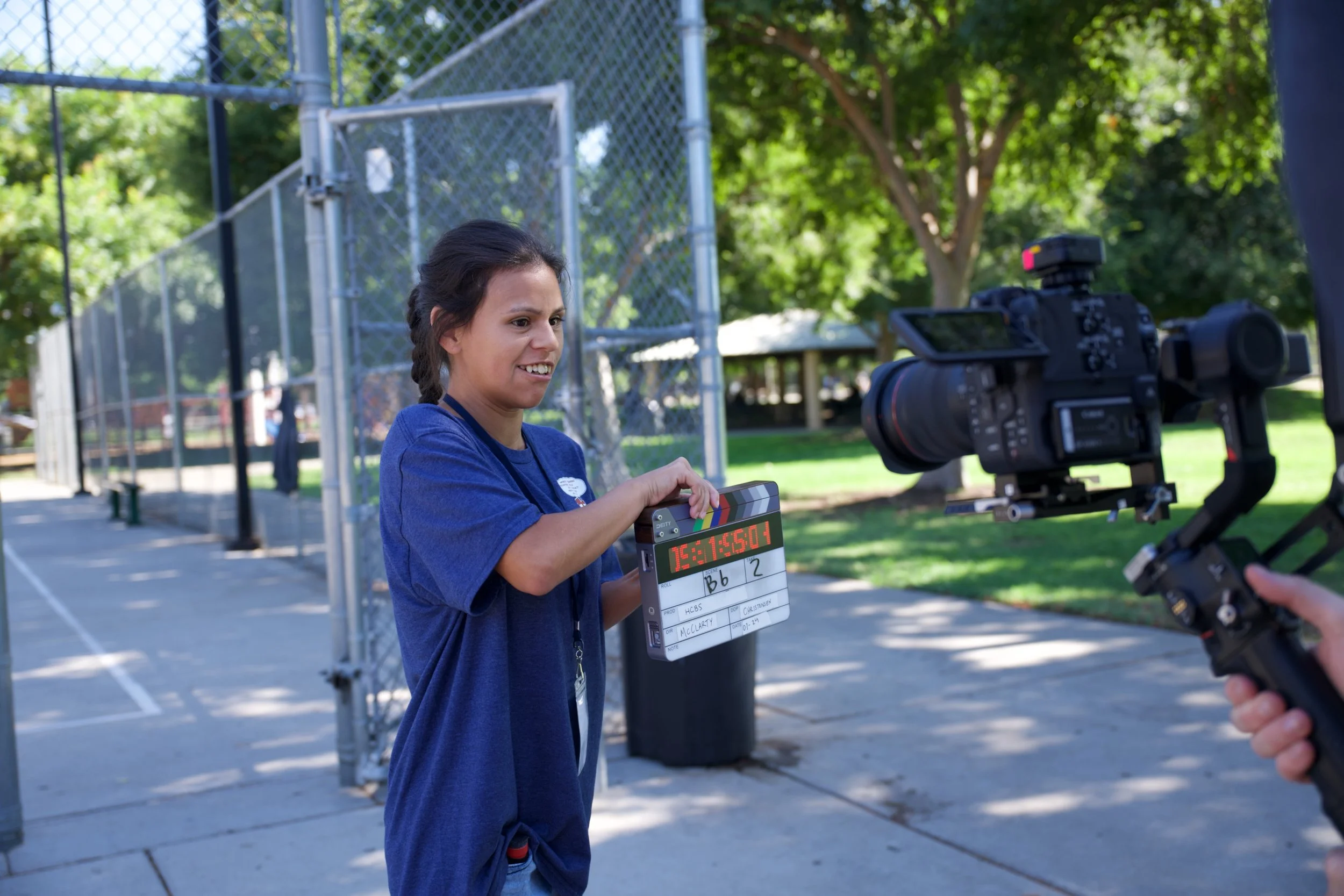 A young girl holding a clapperboard during an outdoor video shoot in a park on a sunny day, with a professional camera filming her.