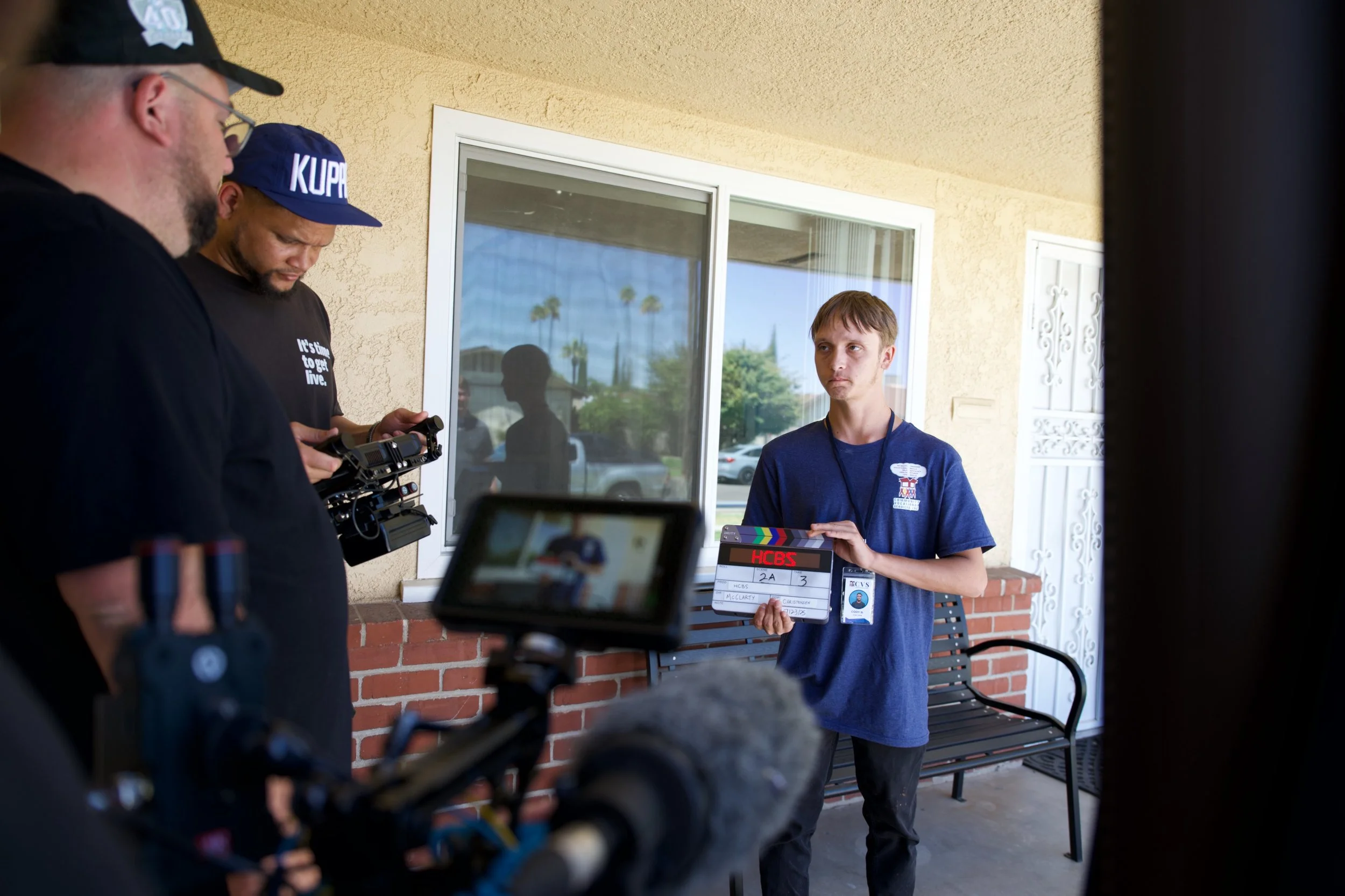 Young man holding a film slate during a video shoot on a porch with two crew members filming.