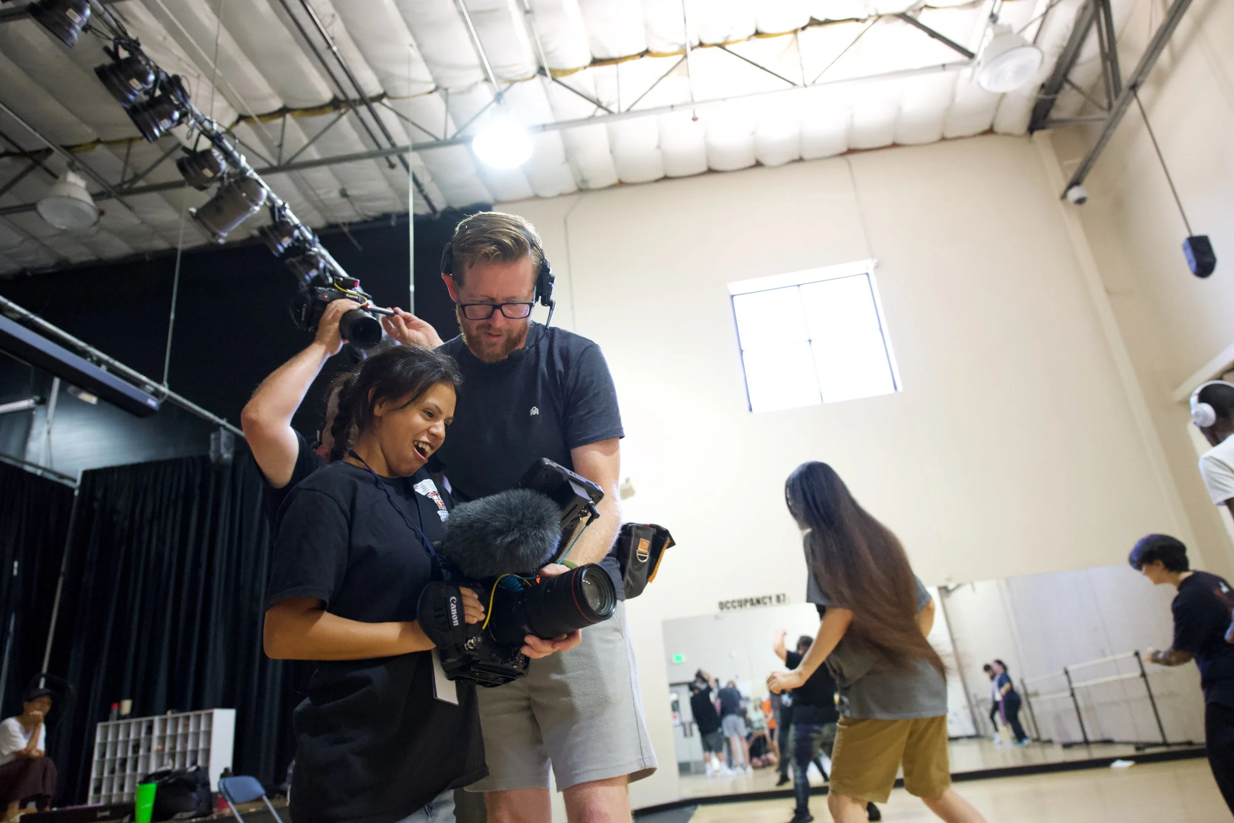 Two people, a woman and a man, are working together inside a large, well-lit gym or studio. The woman is holding a professional camera with a microphone, looking at its screen, while the man is standing behind her, wearing headphones and looking at t