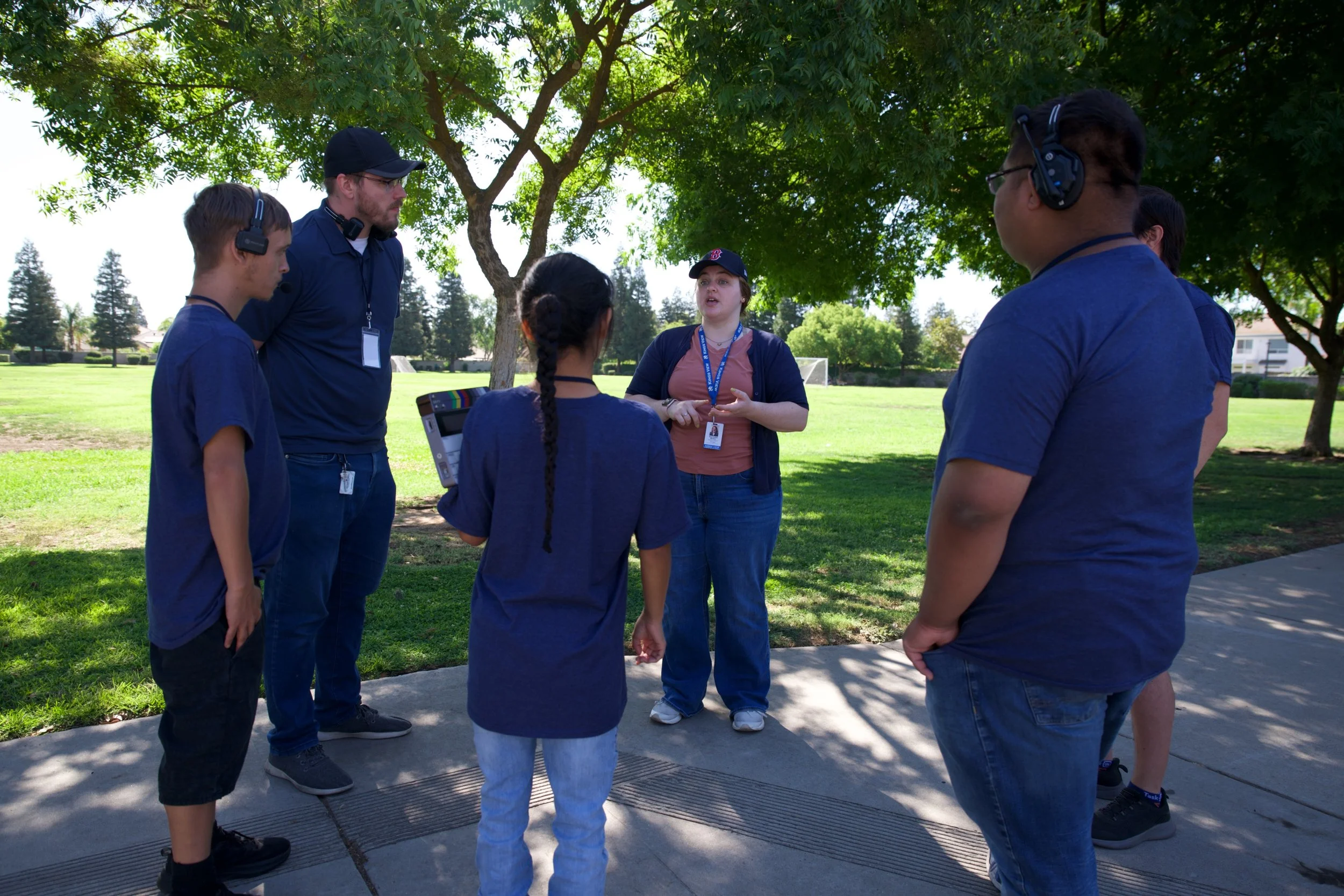 A group of six diverse people, including children and adults, gathered outdoors under trees, listening to a woman speaking. Some are wearing headsets, and one person is holding a booklet or tablet. They appear to be participating in an outdoor educat